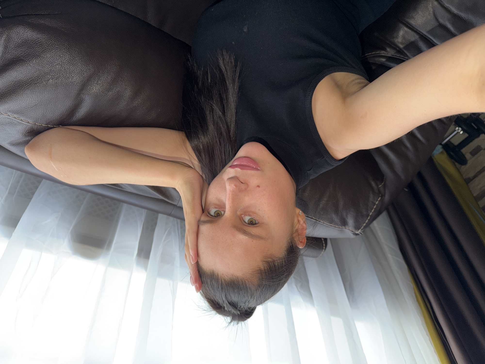 The image features a young woman lying on her back on a dark brown leather sofa, taking a selfie from an upside-down perspective. She has dark hair, which appears to be pulled back or swept away from her face, and is wearing a black ribbed top with a high neckline. Her eyes are light-colored (appearing green or hazel) and gaze directly into the camera. Her lips are slightly parted, showing a glossy finish. One arm is extended, likely holding the camera, while the other arm is bent with her hand gently touching the side of her head.
The background reveals a window with bright light streaming through, covered by sheer white curtains. To the right, a section of darker, possibly brown or olive-green, curtains is visible. The setting is indoors, consistent with a home environment such as a living room or bedroom, situated in Khimki, Russia. The bright illumination from the window indicates that the photo was taken during the daytime. The woman's expression is neutral and direct, suggesting she is posing for the photograph. There is no visible text in the image.