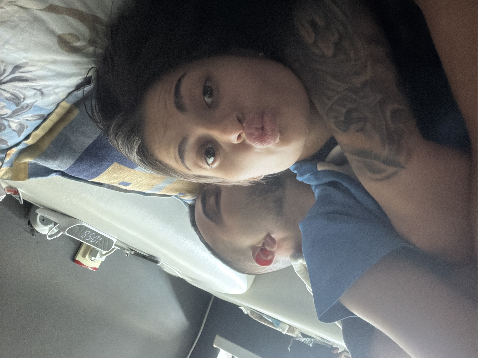 A selfie depicts a woman and a man lying down on a bed in a room. The woman, positioned at the top of the frame, has dark hair and is making a kissing face toward the camera. Her left arm is visible, featuring a tattoo. The man is situated below her, with a portion of his face visible. He is wearing a blue shirt. The bed has a pillow with a patterned pillowcase. A digital clock, showing the time 10:58, is mounted on the wall above the bed. The location is identified as Unknown, Unknown.