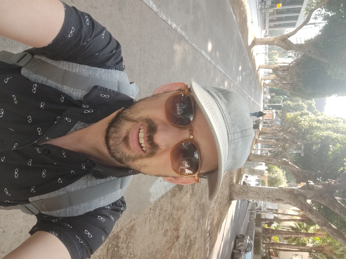 The image depicts a smiling man taking a selfie outdoors in what appears to be Tel-Aviv, Israel. He has a dark beard and is wearing brown-framed sunglasses, a light grey baseball cap, and a black short-sleeved button-up shirt patterned with small white stylized eyeglasses or figure-eights. A grey backpack is visible over his shoulders.
The man's wide smile conveys a cheerful and content emotion. He appears to be enjoying a sunny day, likely during a casual stroll or outing. The background reveals an urban street scene with a wide pedestrian path or sidewalk lined with various trees, some with gnarled trunks and others resembling palm trees. A paved road runs alongside, with parked cars and distant buildings. The bright sunlight and clear conditions suggest a pleasant morning or afternoon. The overall setting is characteristic of a vibrant Mediterranean city. The visible text from the OCR corresponds to the repeating design elements on his shirt rather than coherent words.