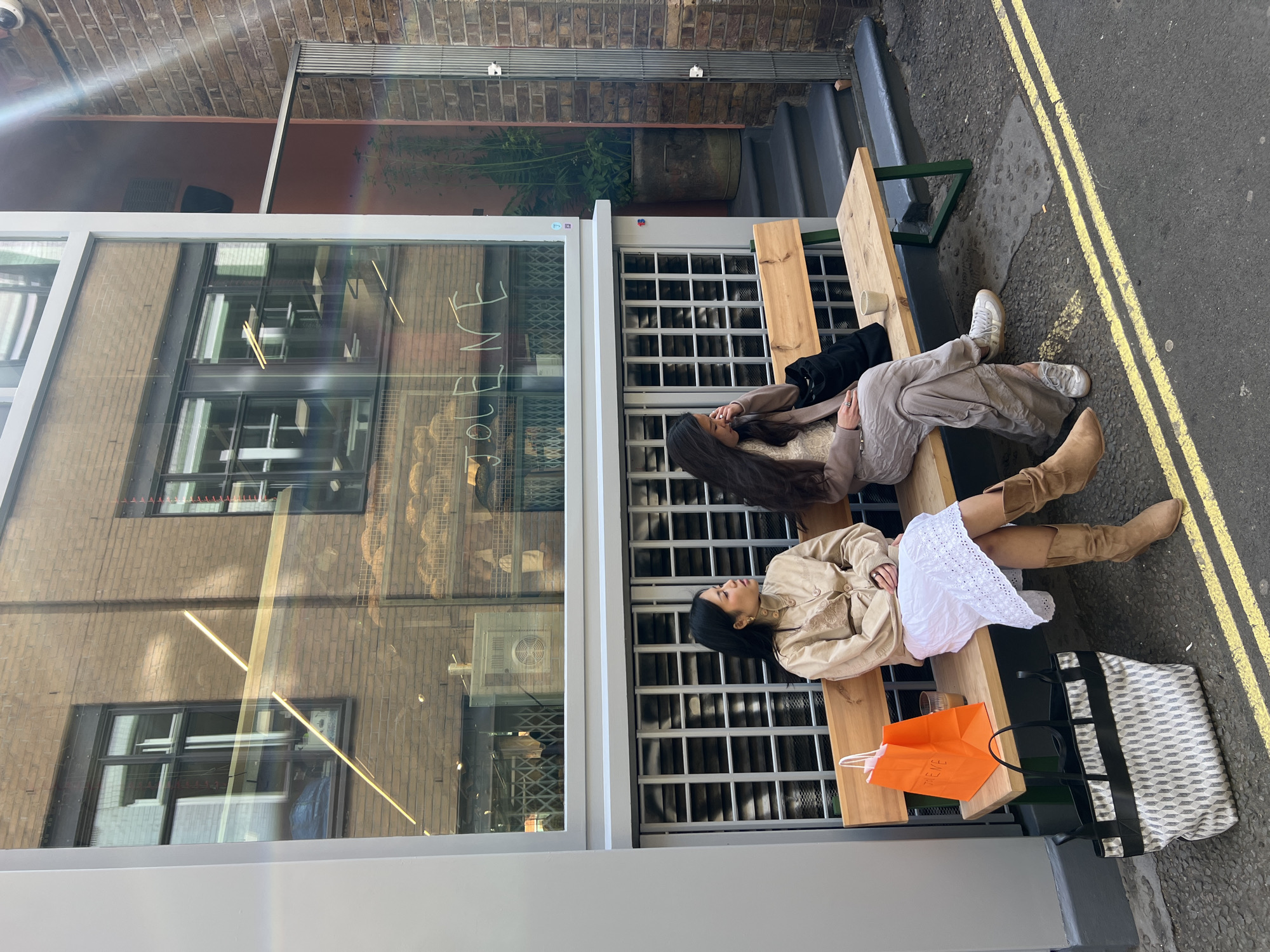 This image, captured from a rotated perspective, depicts two women relaxing on an outdoor wooden bench in what appears to be a London urban setting. The bench is situated outside a modern glass-fronted building, possibly a cafe or shop, and borders a street marked with distinct double yellow lines.
The woman positioned closer to the street is wearing a light beige or brown collared jacket over a white lace skirt, complemented by tall, light brown boots. She has dark hair. Beside her on the bench is a bright orange shopping bag displaying the text "TREMB." A large tote bag with a black and white geometric pattern rests on the ground near her, and a clear glass or cup is on the bench.
The second woman, seated closer to the building, is dressed in a light brown top, loose-fitting light-colored pants, and white sneakers. She also has long dark hair and appears to be looking upwards or slightly away. A black bag is placed next to her on the bench.
The glass facade of the building reflects the brick architecture of the opposing structures and glimpses of the interior, which seems to contain shelves, possibly displaying baked goods. Partially visible, reflected text on the window pane includes what might be "FINE." Above the building, further brickwork is visible, and a security camera is mounted. A large potted plant can be seen in a recess further along the alleyway.
The scene unfolds during daytime, indicated by the clear lighting and visible sunlight, which casts reflections and potentially a sunbeam. The double yellow lines and classic brick buildings are strong cues pointing to a location in the United Kingdom, specifically London. The women appear to be taking a relaxed break from their activities.