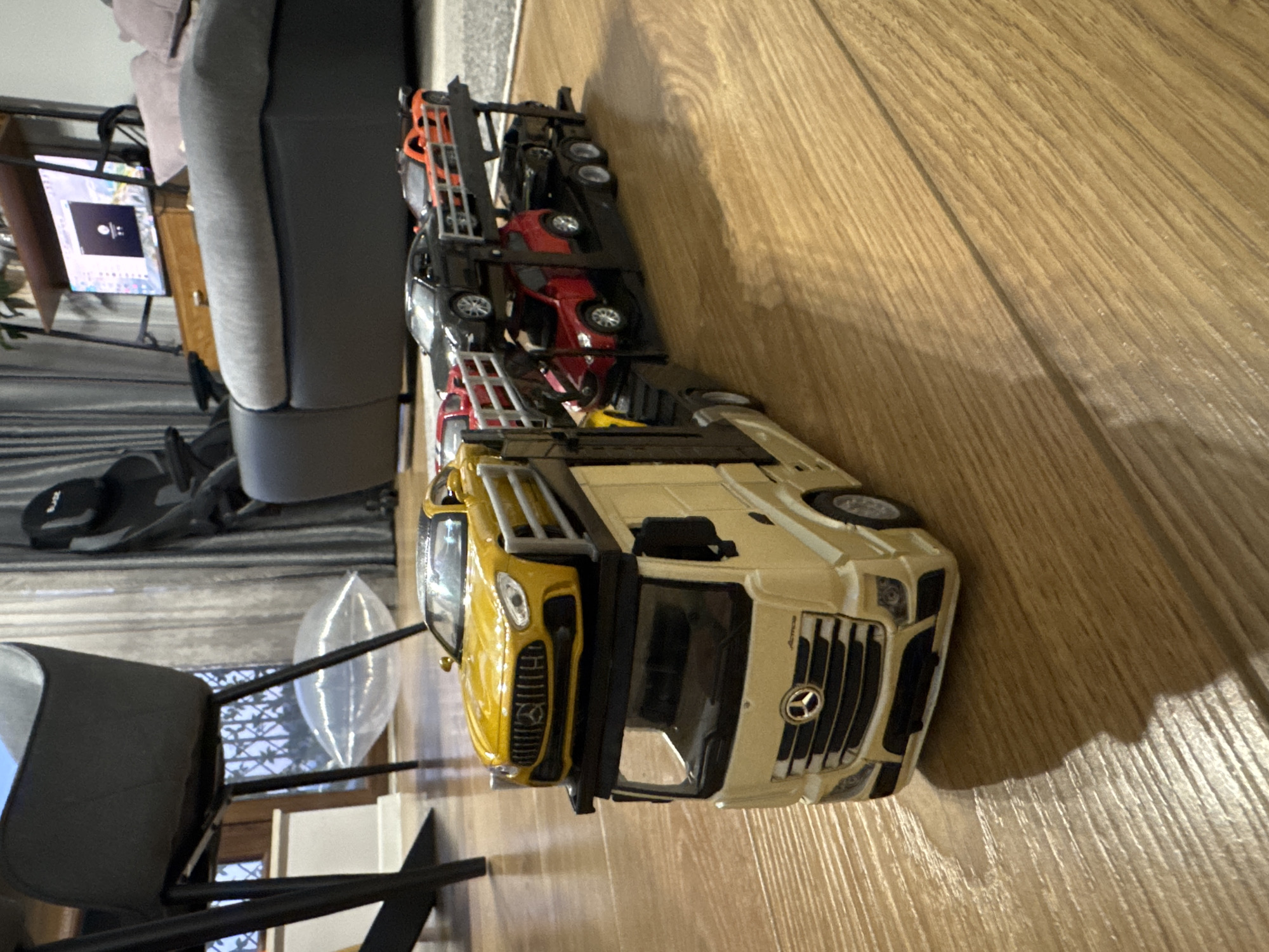 The image shows a toy car carrier truck with multiple toy cars on top, sitting on a wooden floor. The truck is primarily beige and yellow. The toy cars on the carrier appear to be various colors including red, black, and orange. A chair and a portion of a couch are also visible in the background. The location cannot be ascertained from this image.