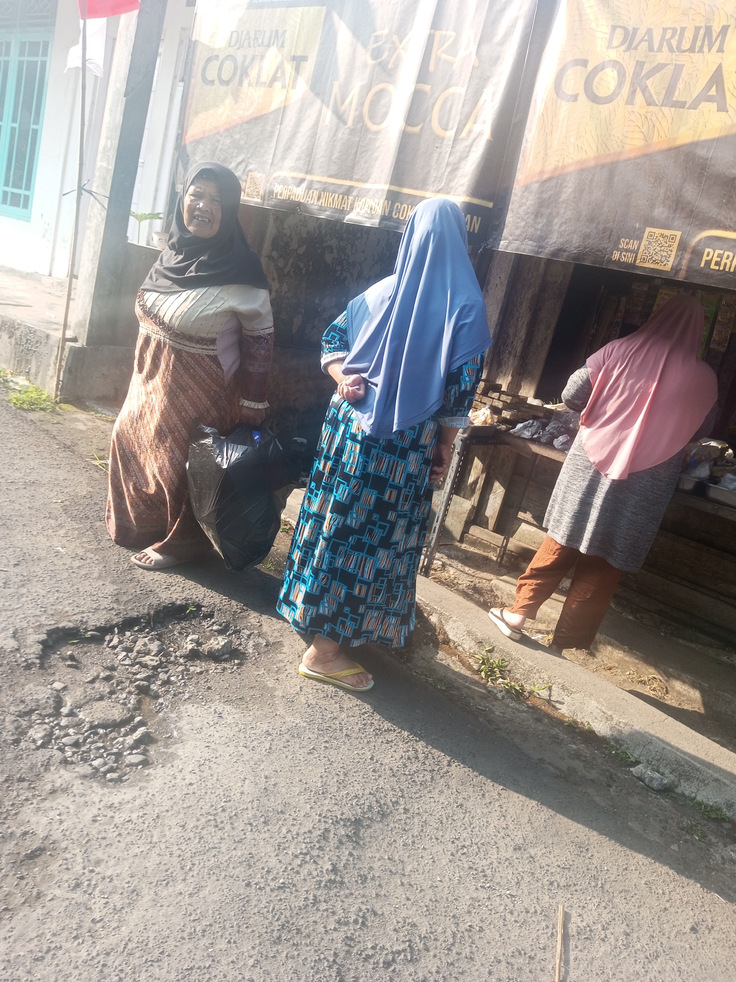 In Krasak, Indonesia, three women stand on a paved road. The woman on the left, wearing a black hijab and a patterned dress, is smiling and holding a black garbage bag. The woman in the middle, dressed in a blue hijab and a blue and black patterned dress, faces away from the camera. The woman on the right wears a pink hijab and a gray tunic with brown pants, and she is also facing away from the camera. Behind them, a banner with the text "DJARUM COKLAT" and a QR code is visible. The environment appears to be outdoors, with a building and a street visible.