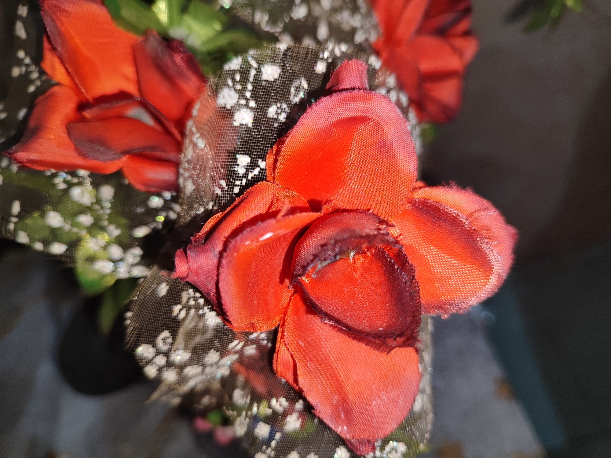 A close-up, shallow depth of field photograph captures artificial red flowers. The flowers, made of fabric, are arranged on what appears to be a dark mesh material adorned with small white dots, possibly glitter or embellishments. The lighting creates highlights and shadows on the petals, revealing their textured surface. The background is blurred, with hints of green foliage and dark, out-of-focus shapes. The scene suggests a decorative arrangement, perhaps part of a bouquet or floral display, and is not indicative of any specific location within Bondowoso, Indonesia.