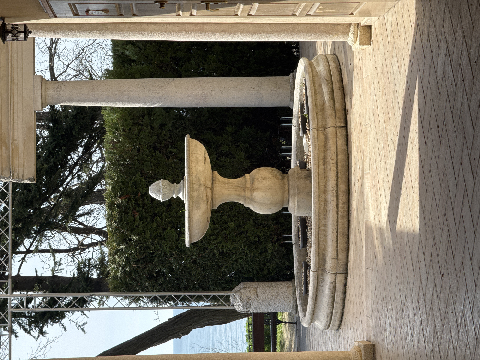 The image captures an outdoor scene, likely a terrace or garden, featuring architectural elements and a natural landscape under a clear, sunny sky. In the foreground, several robust stone pillars frame the view, with one particularly prominent on the left, adorned with an ornate outdoor lantern. The ground is paved with light-colored tiles arranged in a distinct diamond pattern, across which long shadows are cast, suggesting either early morning or late afternoon sun.
A decorative, two-tiered stone fountain stands prominently in the midground. It features a sculpted base, a central column supporting two basins, and a finial at its apex. The lower basin of the fountain appears to contain small stones or gravel and possibly some small planters.
Beyond the architectural foreground, a lush natural environment unfolds. Dense evergreen trees, likely conifers, provide a rich green backdrop, interspersed with the skeletal branches of bare deciduous trees. Through the foliage, a metal truss structure is visible, possibly part of a railing, support, or temporary setup. In the distance, a serene body of water, indicative of a sea or large lake, stretches under the bright sky. Given the location context of Varna, Bulgaria, this is likely the Black Sea.
There are no people or specific activities taking place, giving the scene a tranquil and contemplative atmosphere. No visible text is present in the image.