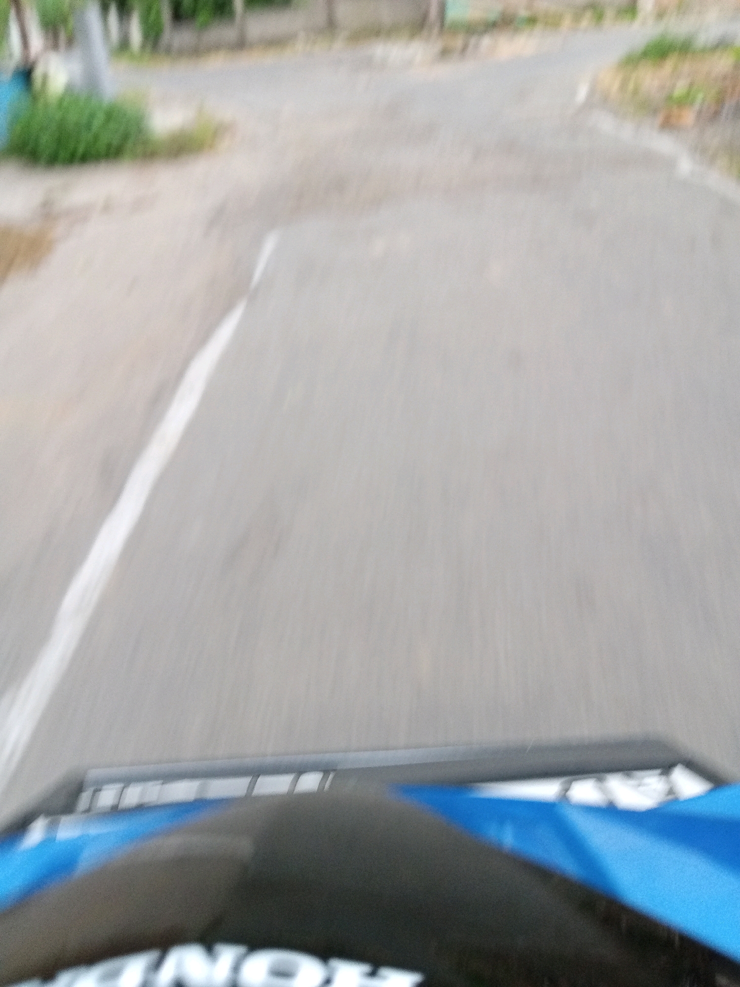 The image is a first-person perspective shot from a moving vehicle, likely a motorcycle, in Bendosari, Indonesia. The foreground shows the upper portion of the vehicle, featuring a dark surface with a blue accent and partially visible white lettering that appears to spell "HONDA". The road ahead is paved, with a white line marking the edge on the left. In the background, there is sparse vegetation, trees, and a blurry view of buildings or structures, suggesting a semi-urban or rural environment. The motion blur indicates the vehicle is in transit.