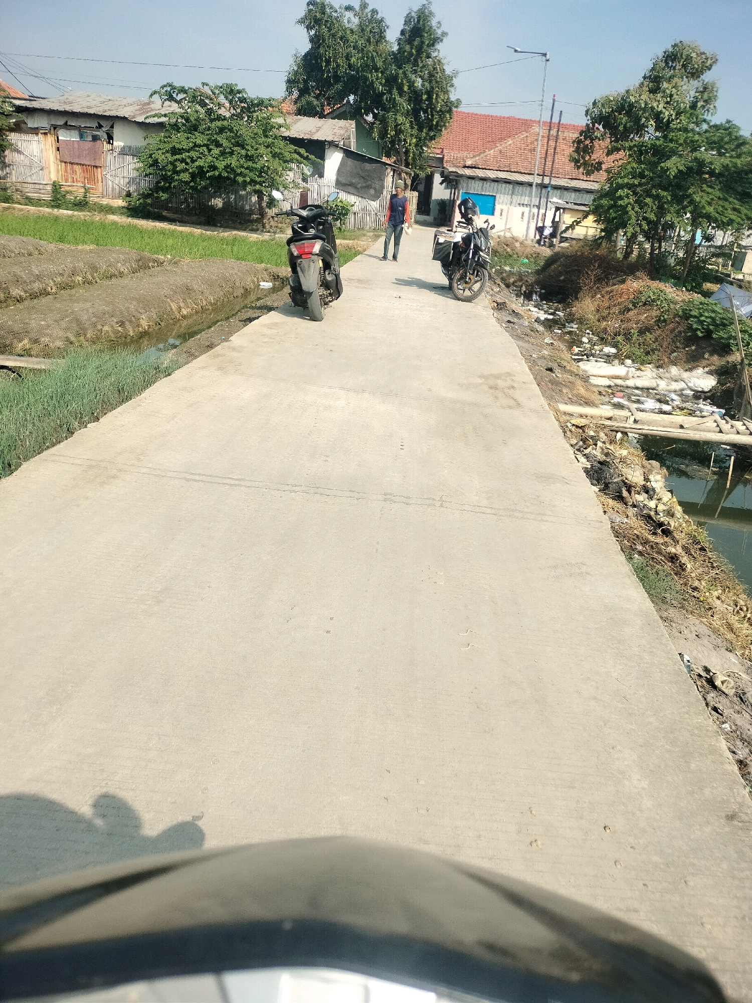 The image depicts a paved road in Mulyasari, Indonesia, with agricultural fields to the left and a canal with debris to the right. Two motorcycles are parked on the side of the road, and a man in a blue shirt and black pants is walking away from the camera. Further down the road, another person is visible, possibly near a motorcycle. The environment suggests a rural or semi-urban area with ongoing development or a lack of waste management. The sky is clear and the lighting indicates it is daytime.