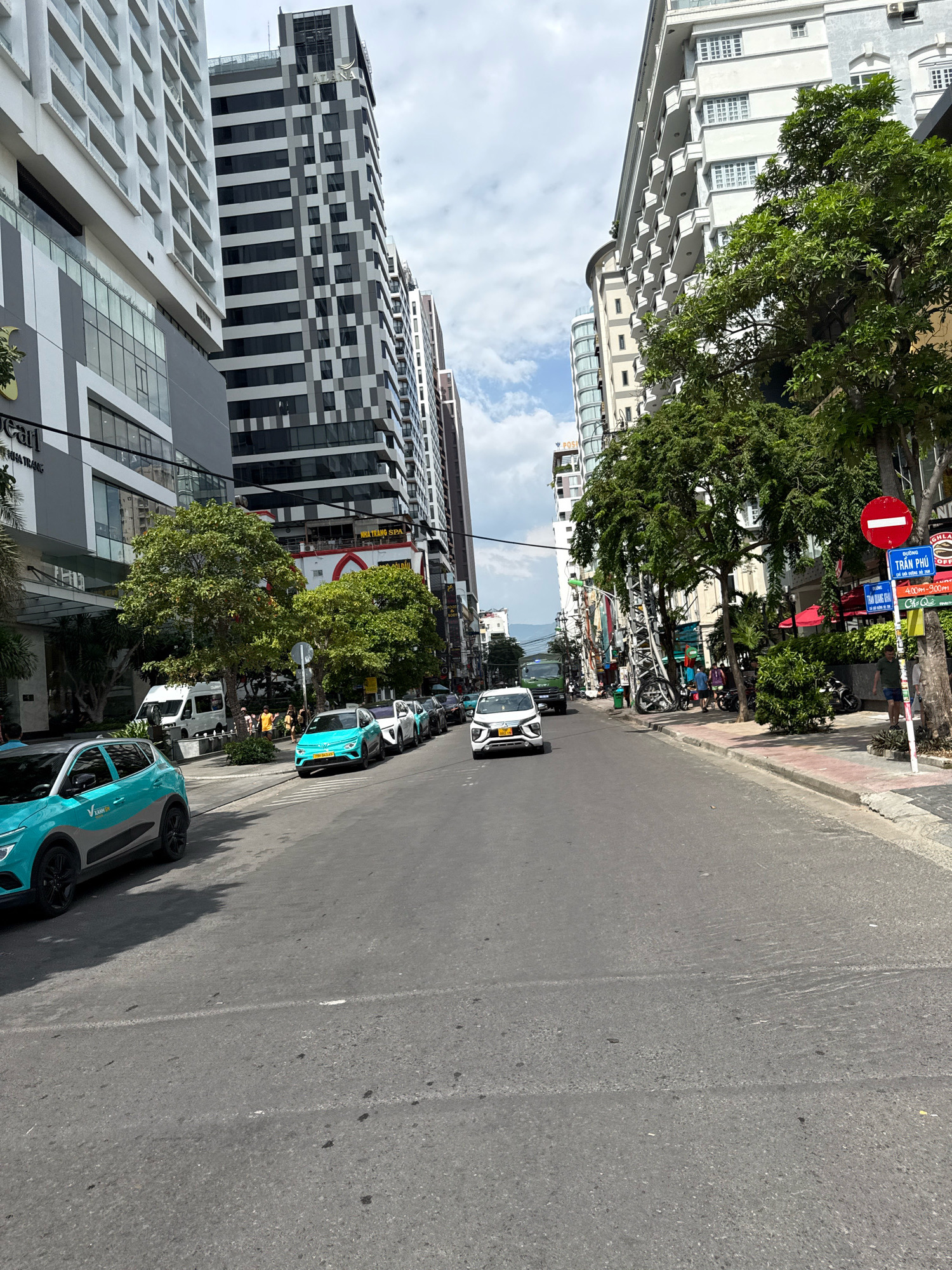 This is a photograph taken from a vehicle, looking down a street in Nha Trang, Vietnam. The street is lined with tall, modern buildings on both sides. On the left, there is a white building with many windows and a darker section with glass facades. Next to it is a tall building with a distinctive black and white checkered pattern. On the right, buildings are also tall with many windows, and large trees with lush green foliage are planted along the sidewalk.
The street itself is asphalt and appears to be a multi-lane road. Several cars are visible: a bright teal car is in the foreground on the left, and a white SUV is in the middle of the road. Further down the street, a line of cars is parked or stopped. Pedestrians are visible on the sidewalks, and some appear to be standing in small groups. There are also traffic signs and streetlights. The sky is partly cloudy, indicating it is daytime. The overall impression is of a busy urban environment in a coastal city.