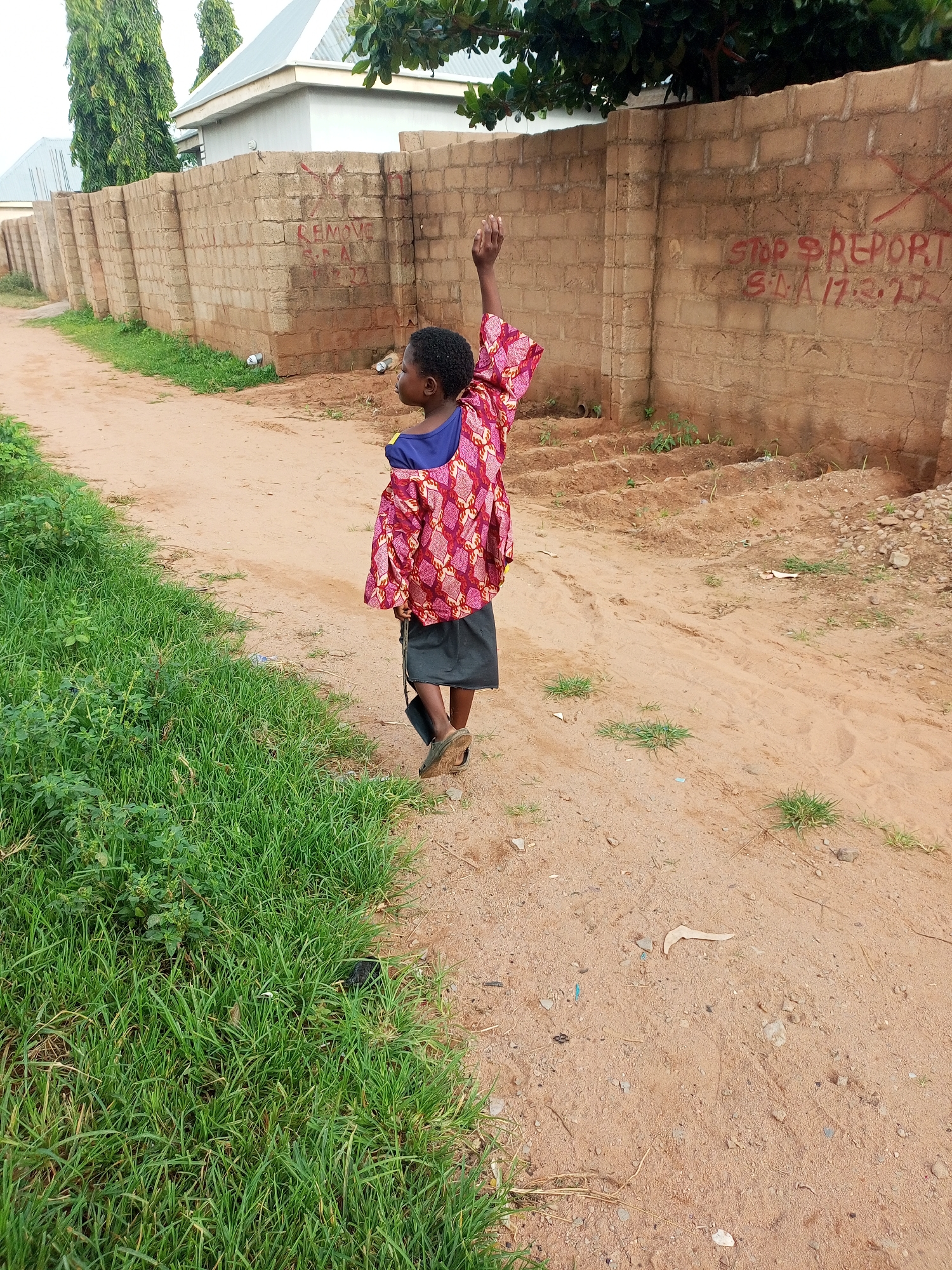 A young child, appearing to be of Nigerian descent, stands on a dirt path in what seems to be a residential area of Tempure, Nigeria. The child is facing away from the camera, looking towards a brick wall. They are wearing a vibrant, patterned shirt and a dark skirt, with sandals on their feet. The child's right arm is raised, as if waving or pointing. The brick wall behind the child has red spray-painted writing on it, with the phrase "STOP REPORT" visible, along with some numbers and letters that are partially obscured. To the left of the path, there is a patch of green grass, and beyond that, some buildings with grey roofs are visible. The ground is a mixture of dirt and sand, with some scattered rocks and dry vegetation. The overall scene suggests a casual, everyday moment in a somewhat rural or less developed urban setting.