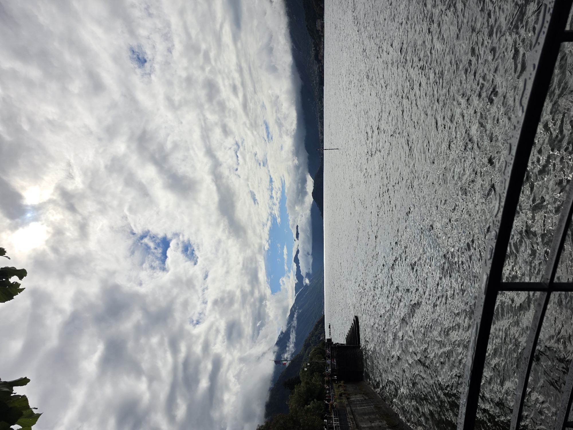 The image depicts a cloudy day over a body of water, likely a lake, with mountains in the background. In the foreground, a black metal railing runs horizontally across the bottom of the frame. To the left, a stone embankment with steps leading down to the water is visible. Several flags are planted on the embankment, and trees line the edge. People are seen walking and standing on the embankment. The water is choppy, with ripples and reflections. In the middle distance, a single post sticks out of the water. The sky is filled with thick, grey clouds, with patches of blue sky visible in the distance. The overall impression is a slightly overcast but scenic view of a lakeside town, potentially San Giovanni, Italy, given the presence of Italian flags and the mountainous backdrop.