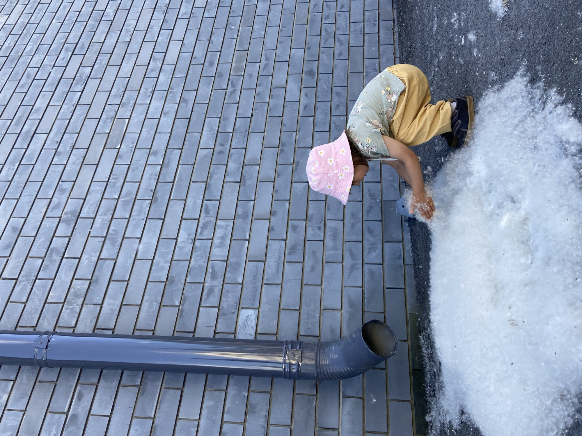 A young child is depicted in a crouching position, interacting with a large accumulation of white, fluffy material on dark asphalt pavement next to a building wall in Tyumen, Russia. The child, observed from a left profile, wears a pink bucket hat adorned with white daisy patterns, a light green short-sleeved top featuring light-colored patterned motifs, and mustard yellow pants. Dark shoes with light brown soles are visible on the child's feet. Both hands are immersed in the white fluff, appearing to scoop or gather it.
The background consists of a uniformly dark grey brick wall, constructed with stretcher bond masonry and dark mortar lines. A dark grey metal downspout runs vertically along the wall, secured by two visible clamps. The downspout bends outwards and downwards near the bottom, ending above the ground where the white fluff has collected. The ground surface is dark asphalt, covered extensively by the white, fibrous material, which resembles poplar fluff. The scene is outdoors, under daylight conditions.