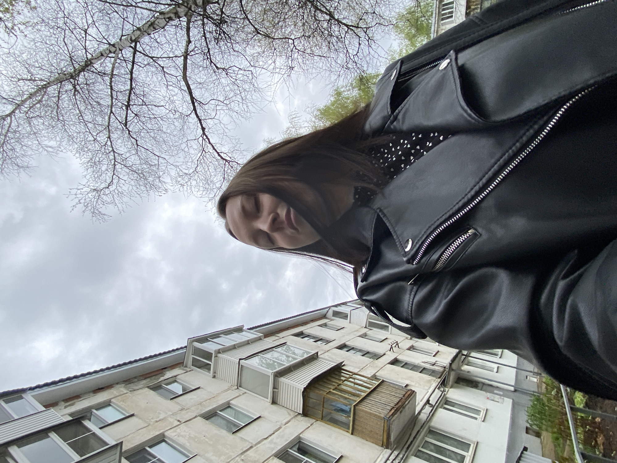 The image, taken from a low angle, features a woman in a black leather jacket, with a building and bare tree branches in the background. The building appears to be an older, multi-story residential structure with many windows, some with enclosed balconies, and a weathered facade. Above the building, tree branches extend into the cloudy sky. Based on the available information, it is impossible to determine the precise location in the city of Chaikovsky, Russia.