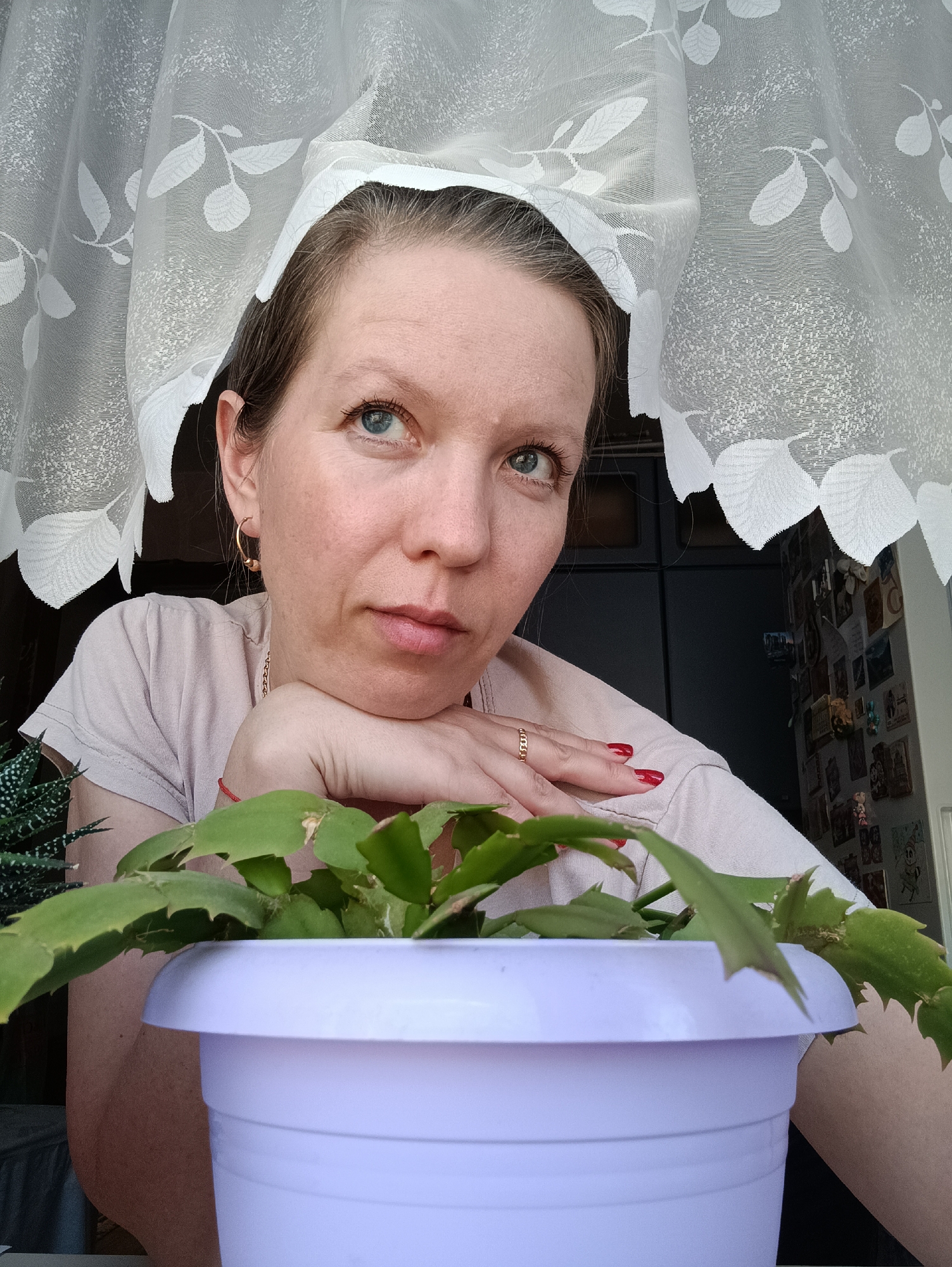 A medium shot captures a woman, potentially in her late 30s or early 40s, resting her head on her hand and looking slightly upwards with a thoughtful expression. She is wearing a light-colored t-shirt and a gold ring is visible on her finger. Her nails are painted red.
A potted plant with green foliage occupies the foreground, slightly obscuring part of her body. The pot is a light purple color.
A white, sheer curtain with a leaf pattern hangs behind her. Behind the curtain, a glimpse of a dark-colored object (possibly a refrigerator or cabinet) is visible on the left, and a wall covered with pictures or postcards can be seen on the right.
The scene appears to be indoors, likely in a home setting. There is no specific visual evidence in the image to confirm the location as Chaikovsky, Russia.
