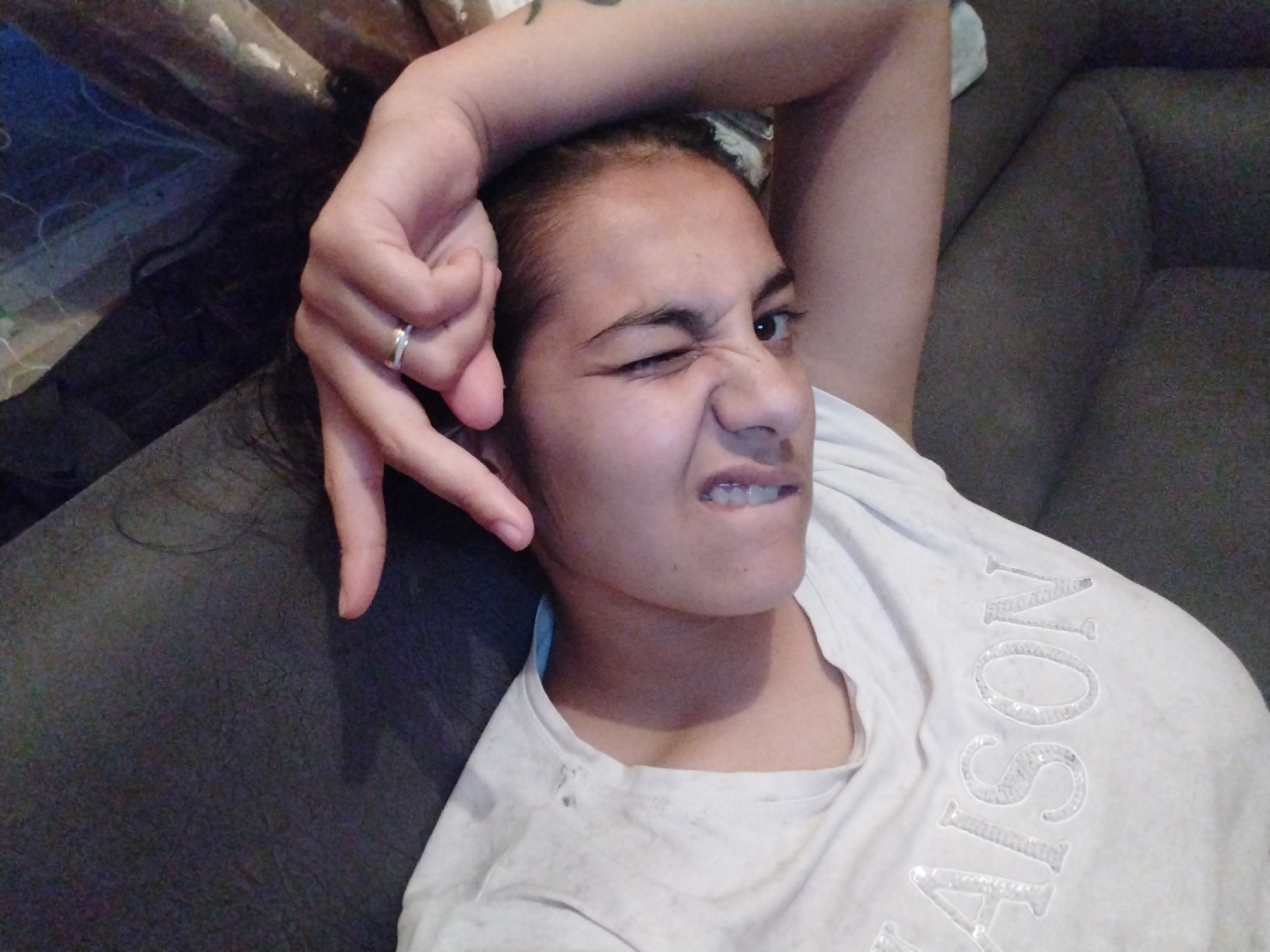A person is lying on a grey couch, winking one eye and making a hand gesture resembling a "V" near their face. They are wearing a light-colored t-shirt with a sequined design. The location is Unknown, Unknown.