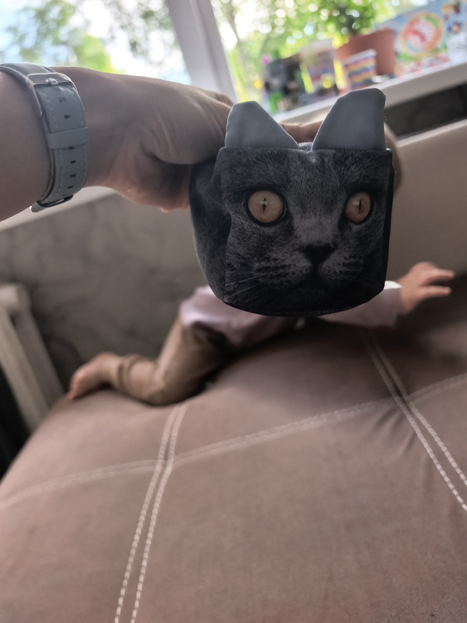 A hand wearing a light blue watch holds a dark grey fabric object with a printed image of a cat's face and ears. The object is positioned in the foreground, obscuring part of a child who is crawling on a brown couch in the background. The child is wearing a light pink long-sleeved shirt and light brown pants. The couch has visible stitching. Sunlight streams in from a window behind the child, illuminating toys on a shelf. The scene appears to be indoors, likely a living room.