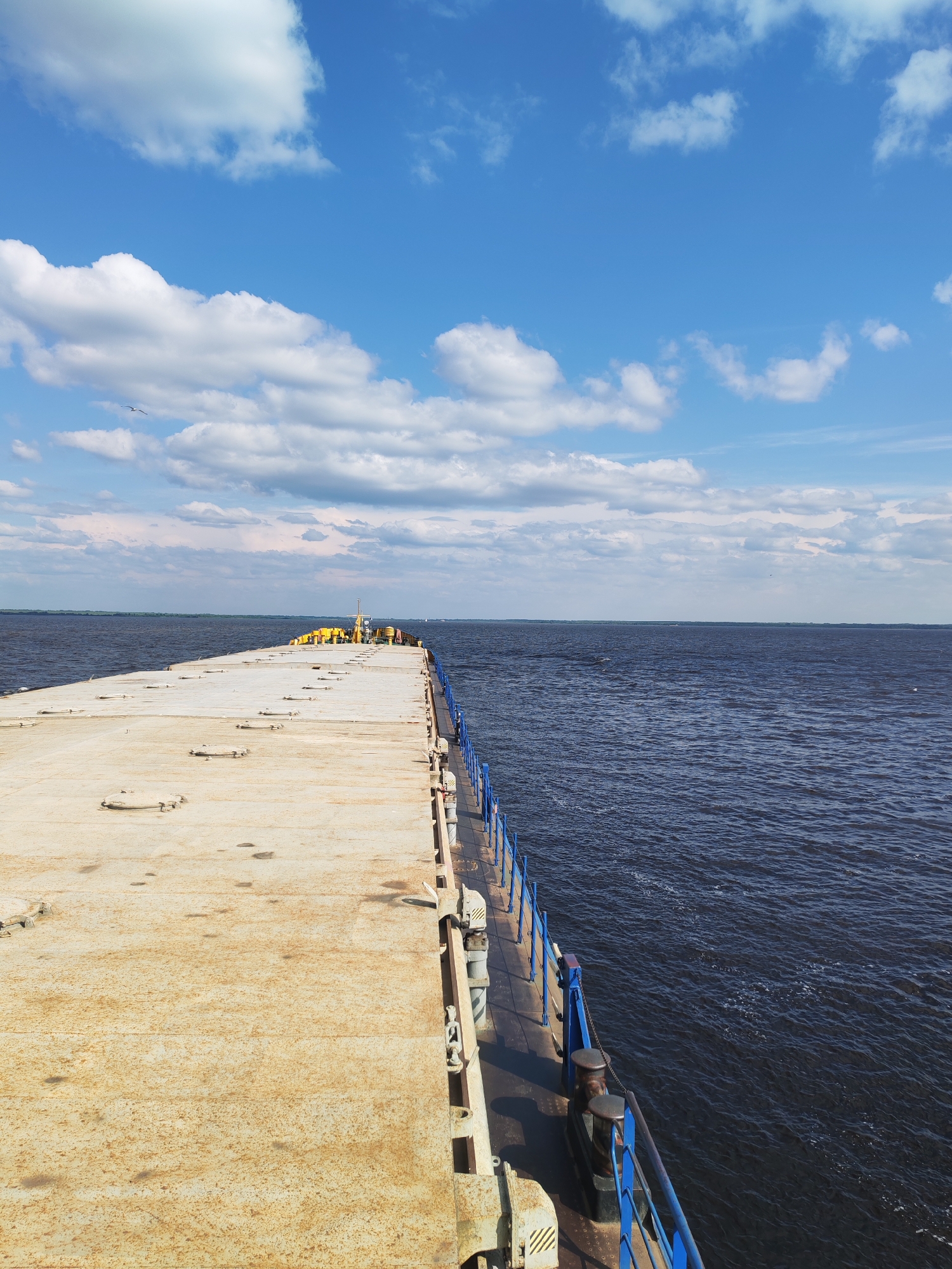 The image depicts a view from the deck of a barge or vessel, looking forward and slightly to the right. The deck is made of a weathered, light-colored material, possibly metal, with visible circular hatches or covers. A blue metal railing runs along the right side of the deck. The dark, rippling water of a wide river or lake extends to the horizon. The sky is bright blue with scattered white cumulus clouds. In the distance, along the horizon, a strip of land is visible, with some structures or lights appearing on the far right. A few yellow objects are clustered on the deck in the distance. The location is identified as Veregonets, Russia.