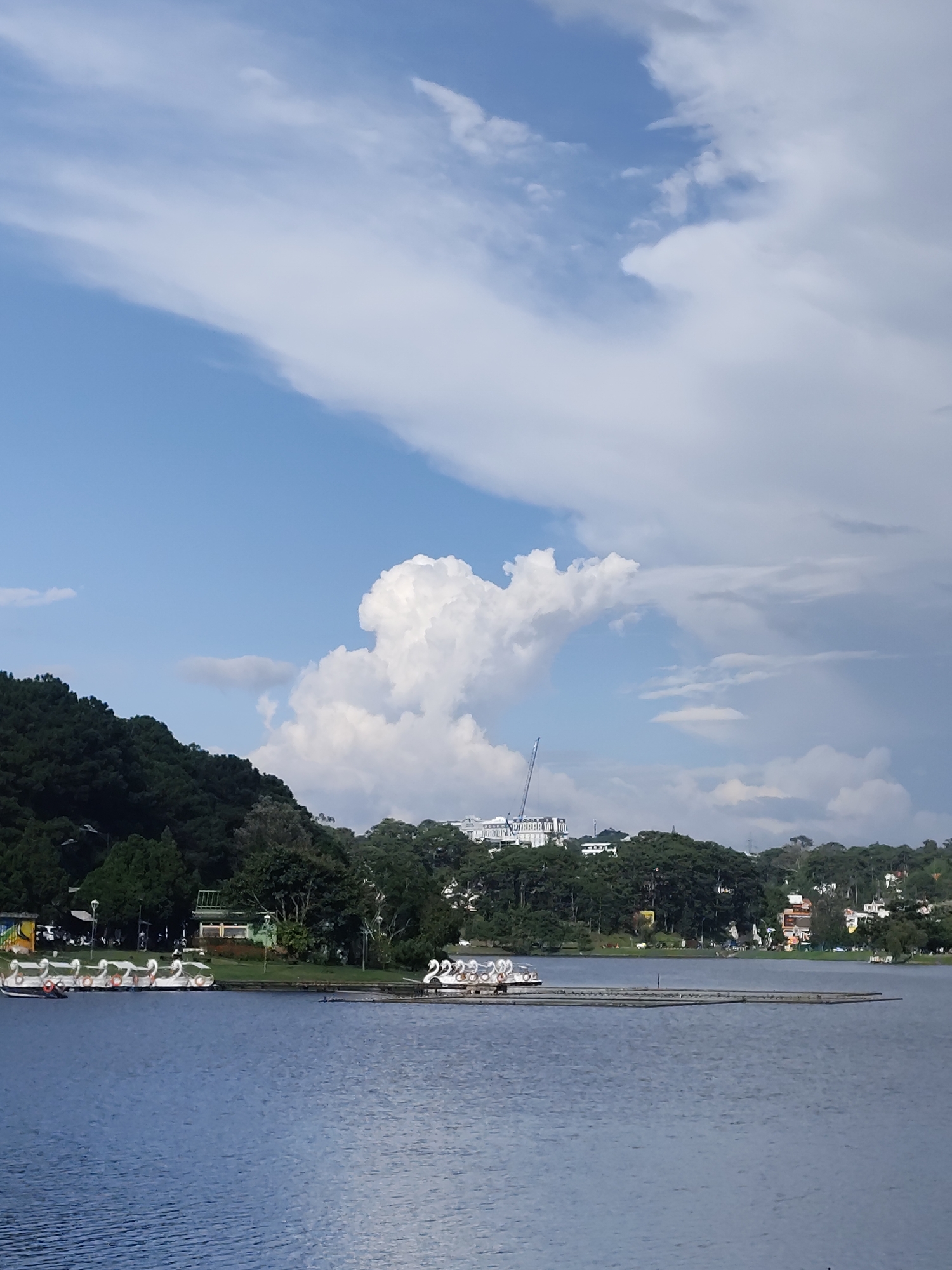 A wide shot captures a serene lake in Da Lat, Vietnam, on a partly cloudy day. The calm, blue water of the lake takes up the lower half of the frame, reflecting the sky. Along the left bank, a lush, dark green forest covers a hillside, with a few buildings and colorful structures visible amidst the trees. In the distance, on a ridge behind the trees, a large white building with a distinctive architectural style can be seen, alongside a construction crane. The sky is a mix of bright blue and large, fluffy white clouds, with some darker, more dramatic clouds towards the right side of the frame. In the foreground of the lake, a floating dock or pier is visible. A line of swan-shaped pedal boats are docked along the left side of the pier, ready for use. Another group of swan boats is further down the pier on the right. The overall atmosphere is peaceful and picturesque, typical of the natural beauty found in Da Lat.