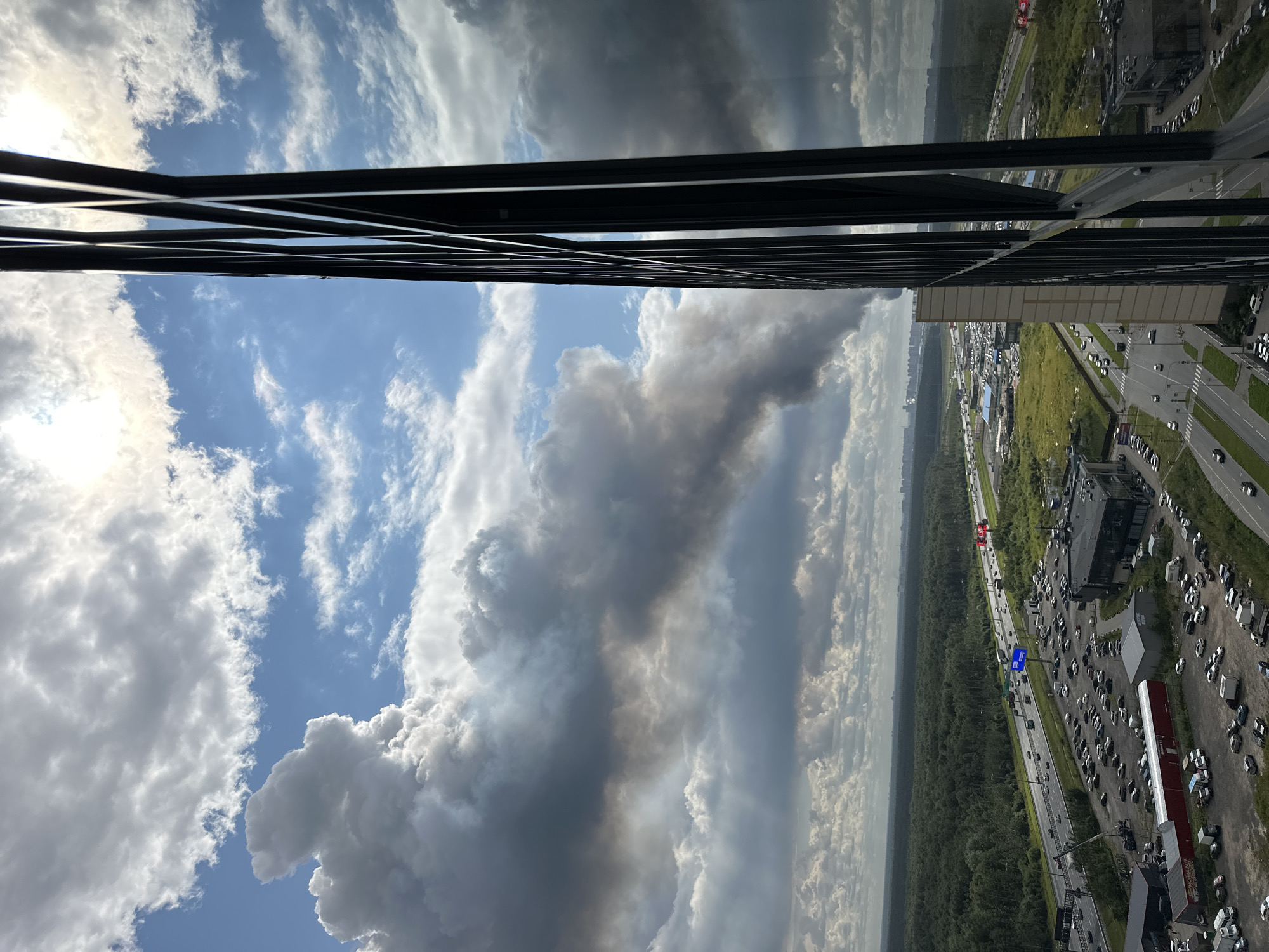 A high-angle, wide shot captures a dramatic sky filled with dark, ominous clouds, partially obscuring the sun. A plume of dark smoke billows across the horizon, suggesting a fire or industrial activity. In the foreground, the sleek, reflective facade of a modern building dominates the right side of the frame, its dark mullions creating a geometric pattern. Below, a wide, multi-lane highway is visible, teeming with vehicles. The surrounding landscape is a dense, green forest that stretches to the horizon, interrupted by urban development and commercial buildings with extensive parking lots filled with cars. The scene appears to be on the outskirts of an urban area, possibly near Novosergievka, Russia, given the context of the city. There are no discernible people in the image.