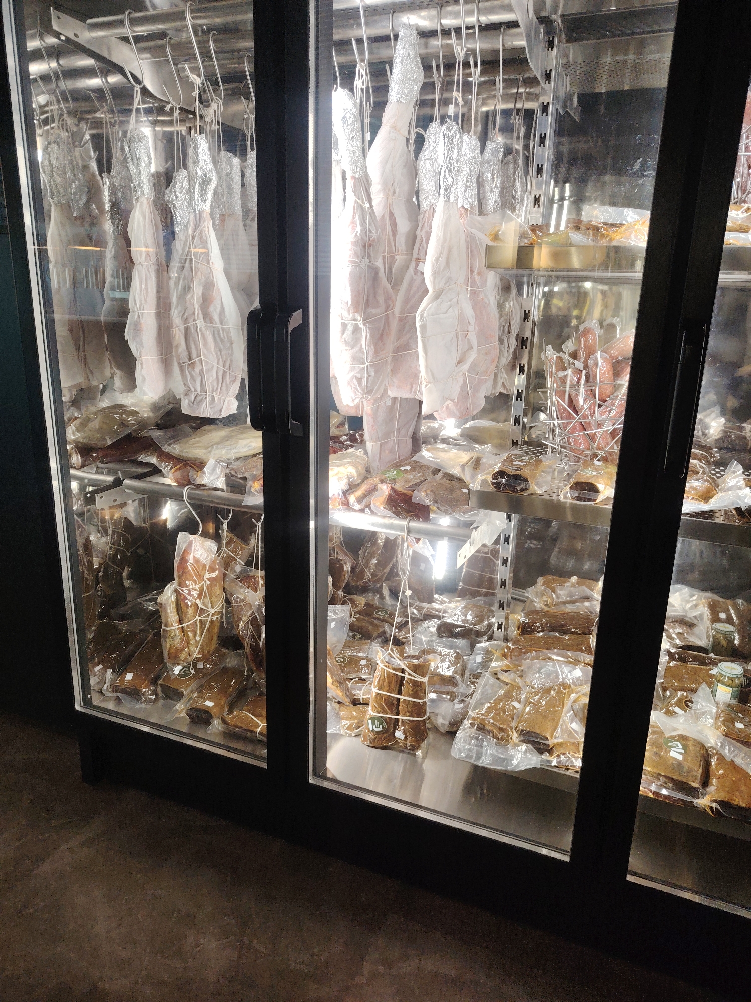 The image shows a refrigerated display case filled with various types of cured meats. The meats are presented in different forms: some are whole and hanging, wrapped in foil and butcher paper, while others are pre-packaged in plastic. The display case has glass doors, revealing multiple shelves stocked with these products. The environment appears to be an indoor commercial space, possibly a deli or a specialty food store. The floor is a dark, textured surface, and the lighting suggests an interior setting. The location is identified as Konigil, Uzbekistan.