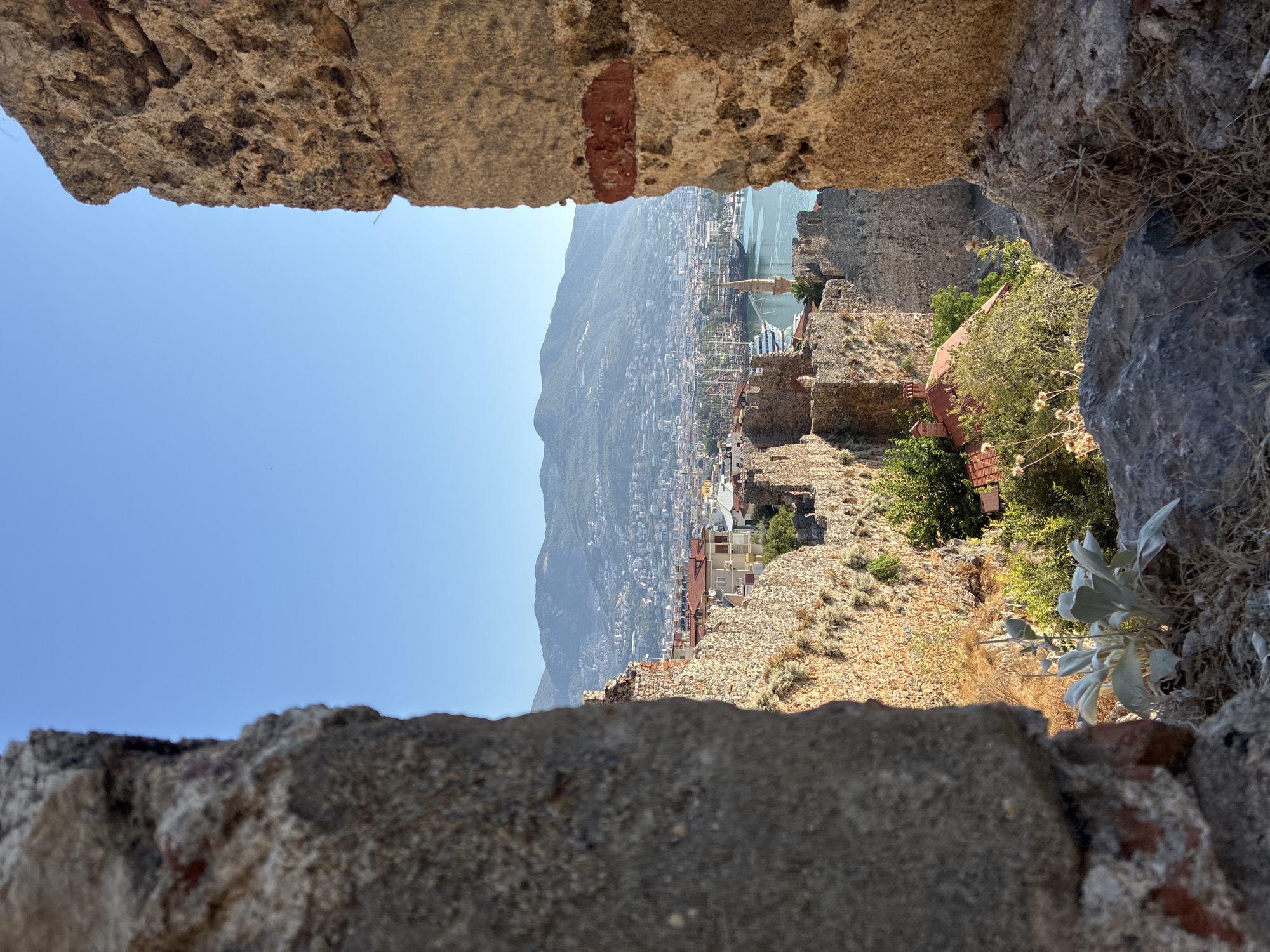 A sun-drenched, medium shot captures a view of Alanya, Turkey, framed by ancient stone walls. The foreground is dominated by rough-hewn stone structures, with a large, weathered rock on the left and a segmented, textured wall on the right, casting shadows and creating a sense of depth. Beyond these fortifications, the city unfolds. Buildings with red-tiled roofs are clustered together, interspersed with lush green trees. In the distance, the azure water of a harbor teems with various vessels, including several tall ships and a white passenger boat. A minaret, characteristic of Turkish architecture, rises elegantly from the waterfront. The backdrop is a range of hazy, blue-green mountains that stretch towards a clear, bright blue sky. The overall impression is one of historical grandeur and a vibrant coastal city bathed in natural light.