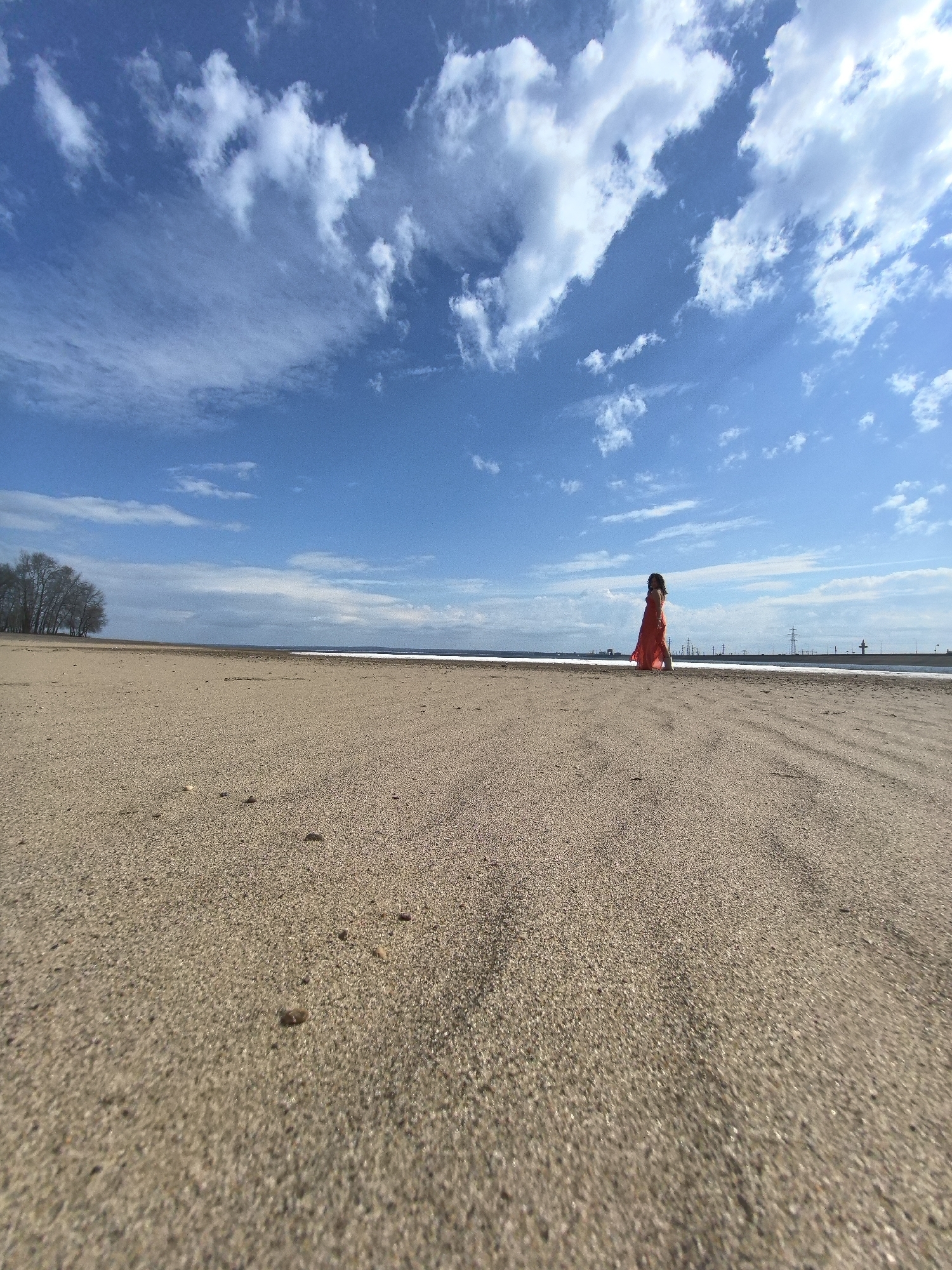 The image presents a wide, low-angle perspective of a vast, sandy beach or shoreline under a clear blue sky. Dominating the foreground is the textured, light brown sand, sparsely dotted with small pebbles, captured from a ground-level viewpoint.
In the middle-right of the frame, a lone individual, possibly a child, is seated or crouching on the sand, wearing a distinctive, bright orange or red outfit. The figure is positioned to appear small against the expansive landscape.
The horizon features a calm body of water, possibly a reservoir or large river, stretching across the mid-ground. On the far left, a cluster of bare-branched trees is visible along the distant shore. To the right, faint industrial structures or power lines can be seen on the distant horizon line.
The scene is illuminated by bright daylight, casting subtle shadows and indicating sunny weather with scattered white cumulus and cirrus clouds in the sky. The low camera angle emphasizes the immense open space and the solitude of the moment. No text is visible in the image. Given the location context of Chaikovsky, Russia, this is likely a view along the Votkinsk Reservoir.