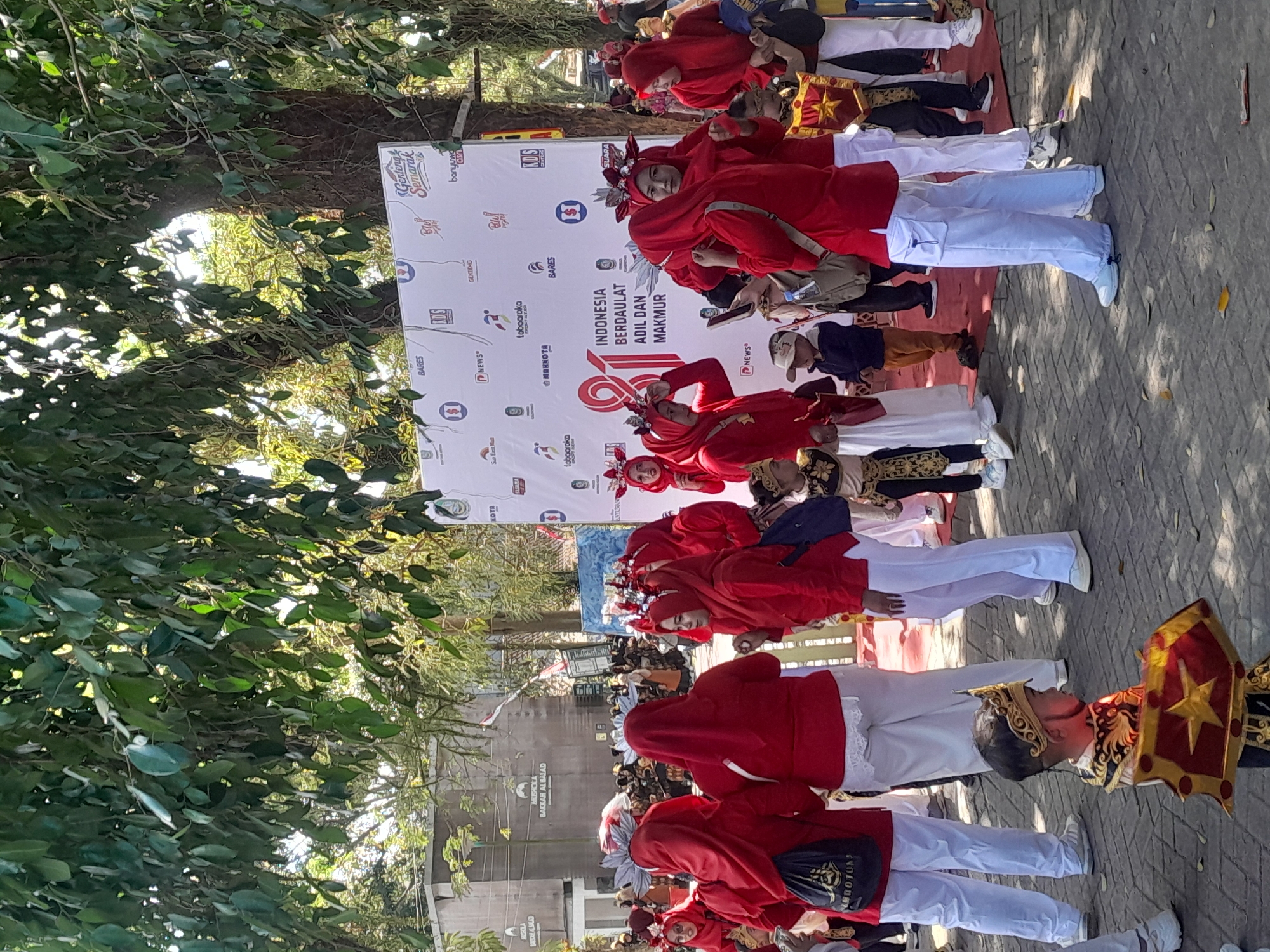In the city of Genteng Kulon, Indonesia, a group of people are gathered outdoors for an event. The scene is dominated by a large white banner featuring the Indonesian flag and text that reads "INDONESIA BERDAULAT ADIL DAN MAKMUR". Numerous individuals, predominantly women and children, are dressed in vibrant red attire, many with matching headscarves and ornate headpieces. Some individuals are wearing white pants, while others are in traditional clothing. A young boy in the foreground is dressed in a golden costume with a shield. The background reveals trees, buildings, and other attendees. The atmosphere appears festive and communal, suggesting a cultural celebration or a public gathering.
