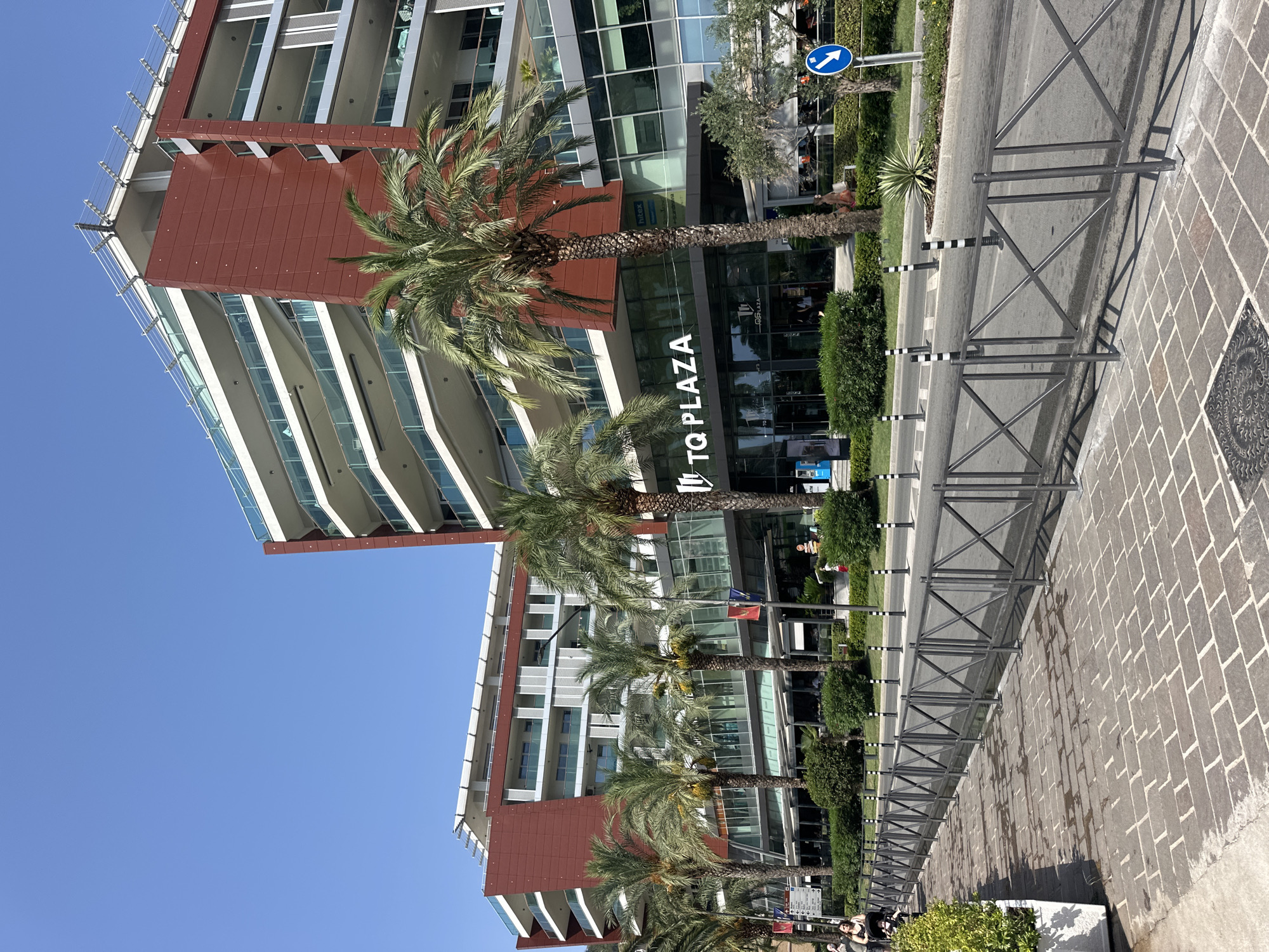 The image shows a modern, multi-story building with a distinctive red facade, a part of the TQ Plaza complex, located in Budva, Montenegro. The building features balconies with glass railings and a curved design. In front of the building, several palm trees are visible, casting shadows on the sidewalk. A street runs alongside the building, separated from the pedestrian walkway by a metal fence. The sidewalk is paved with stone tiles, and there are some small potted plants and streetlights. The sky is clear and blue, indicating a sunny day. A person is visible on the sidewalk, pushing a stroller. Another person is seen walking further down the street. A blue circular sign with a white arrow pointing to the right is also visible on the side of the road. The scene depicts an urban environment, likely a commercial or residential area, with a clear emphasis on contemporary architecture and landscaping.