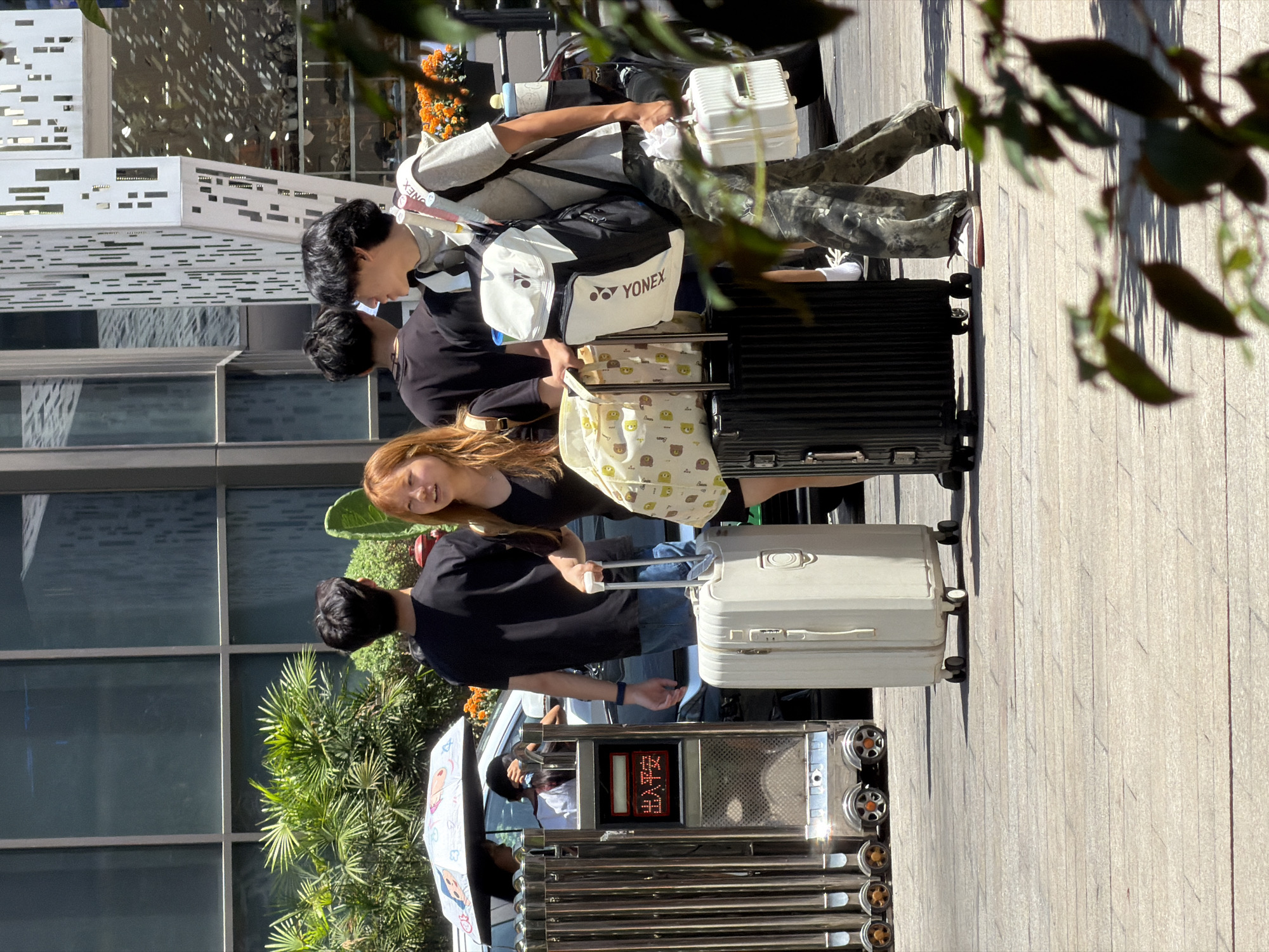 In Yuzhong District, China, on a paved outdoor area with sunlight casting shadows, four young adults are gathered with luggage. Two individuals, one male and one female, stand closer to the viewer, with the male holding the handle of a large, light-colored rolling suitcase, and the female standing next to a black rolling suitcase. The male is wearing a grey t-shirt and camouflage pants, carrying a black and white backpack with "YONEX" branding. The female has reddish-brown hair and is wearing a dark top, with a patterned tote bag placed on top of the black suitcase.
Behind them, two other individuals are visible. A male in a black t-shirt and blue jeans is on the left, with his back to the camera. A second male, also in a black t-shirt, is to the right of the first male, facing slightly away from the viewer.
To the left of the group, a metal gate with wheels is partially visible. A car with an open umbrella, possibly for shade, is behind the gate, with a person sitting in the passenger seat. A sign with red digital characters, appearing to be Chinese, is mounted on the gate structure. In the background, a modern building with large windows and patterned facade elements is seen, along with green foliage and orange flowers.