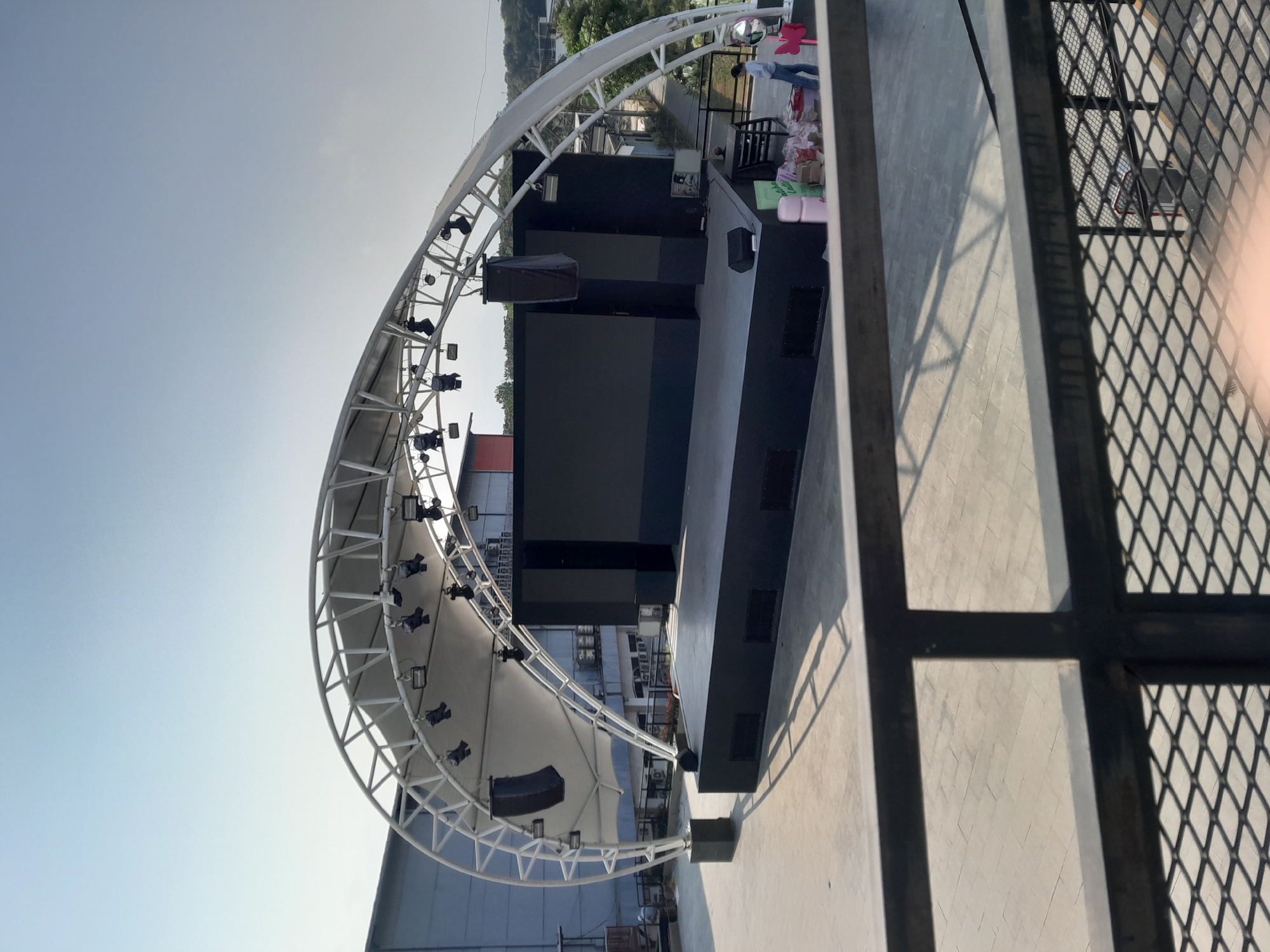 The image depicts an outdoor stage setup under a large, white arched canopy structure. The stage itself is a raised platform with a black surface and what appears to be a large LED screen mounted on the back. Several speakers are visible, attached to the stage and suspended from the canopy. The surrounding area is paved with light-colored tiles, and there is a black metal railing in the foreground. In the background, there are buildings and trees, suggesting an urban or semi-urban environment. A person is standing near the stage, appearing to be organizing some items. The sky is clear and bright, indicating daylight. The scene suggests a venue prepared for an outdoor event or performance in Sempusari, Indonesia.