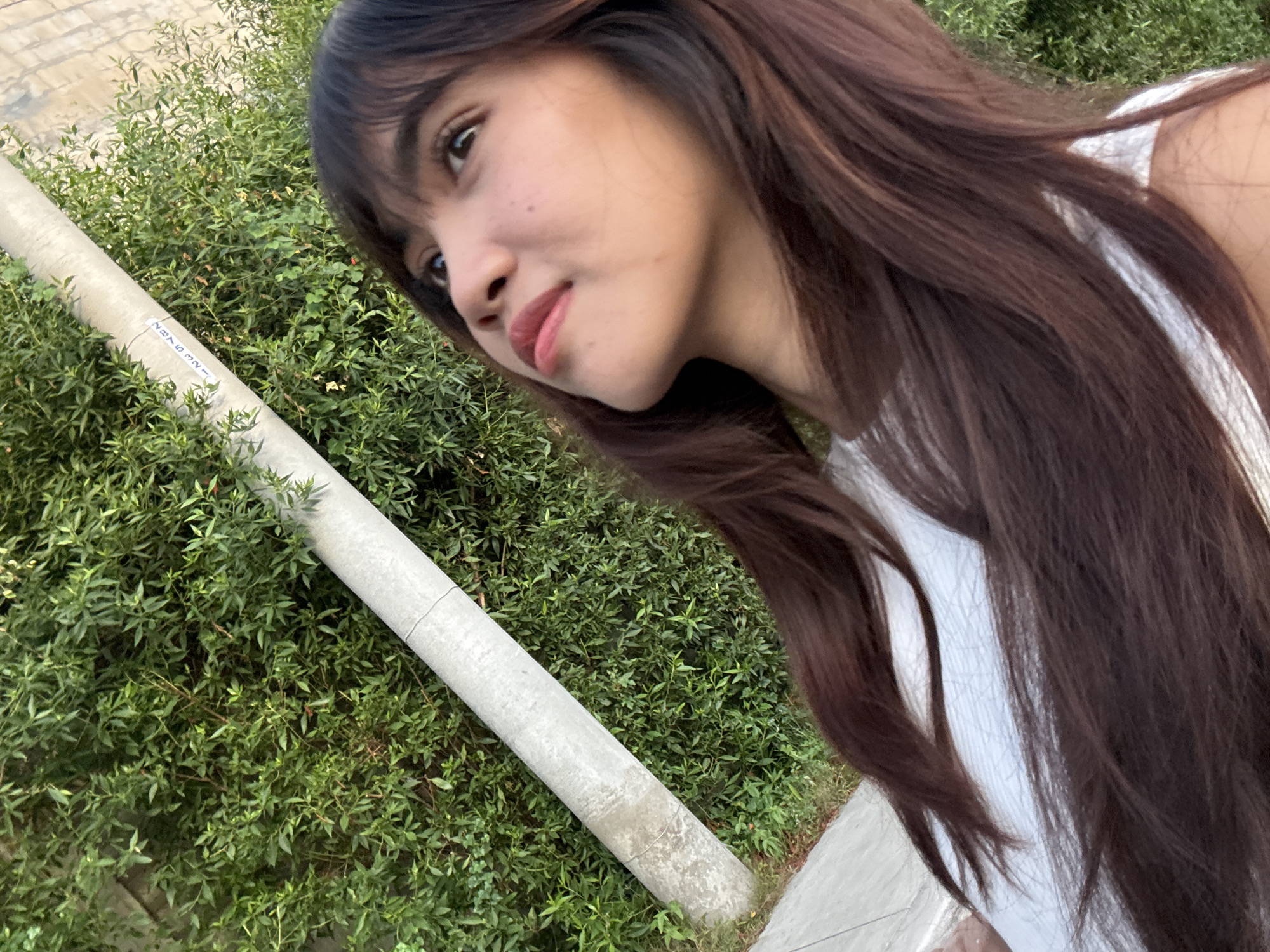 A young woman with long, dark brown hair and bangs is looking upwards and to the right, her head tilted back. She is wearing a white top. Her skin is light brown, and she has full lips and dark eyes. She has a few scattered freckles on her cheek. Behind her, a light gray, cylindrical object, possibly a pole or pipe, is angled diagonally from the bottom left to the top right of the frame. It has some markings or text on it. The background is filled with dense green foliage, suggesting an outdoor setting. The lighting indicates it is daytime, with the sun illuminating the scene from the side. This image does not provide enough visual information to determine a specific location within Davao City, Philippines.
