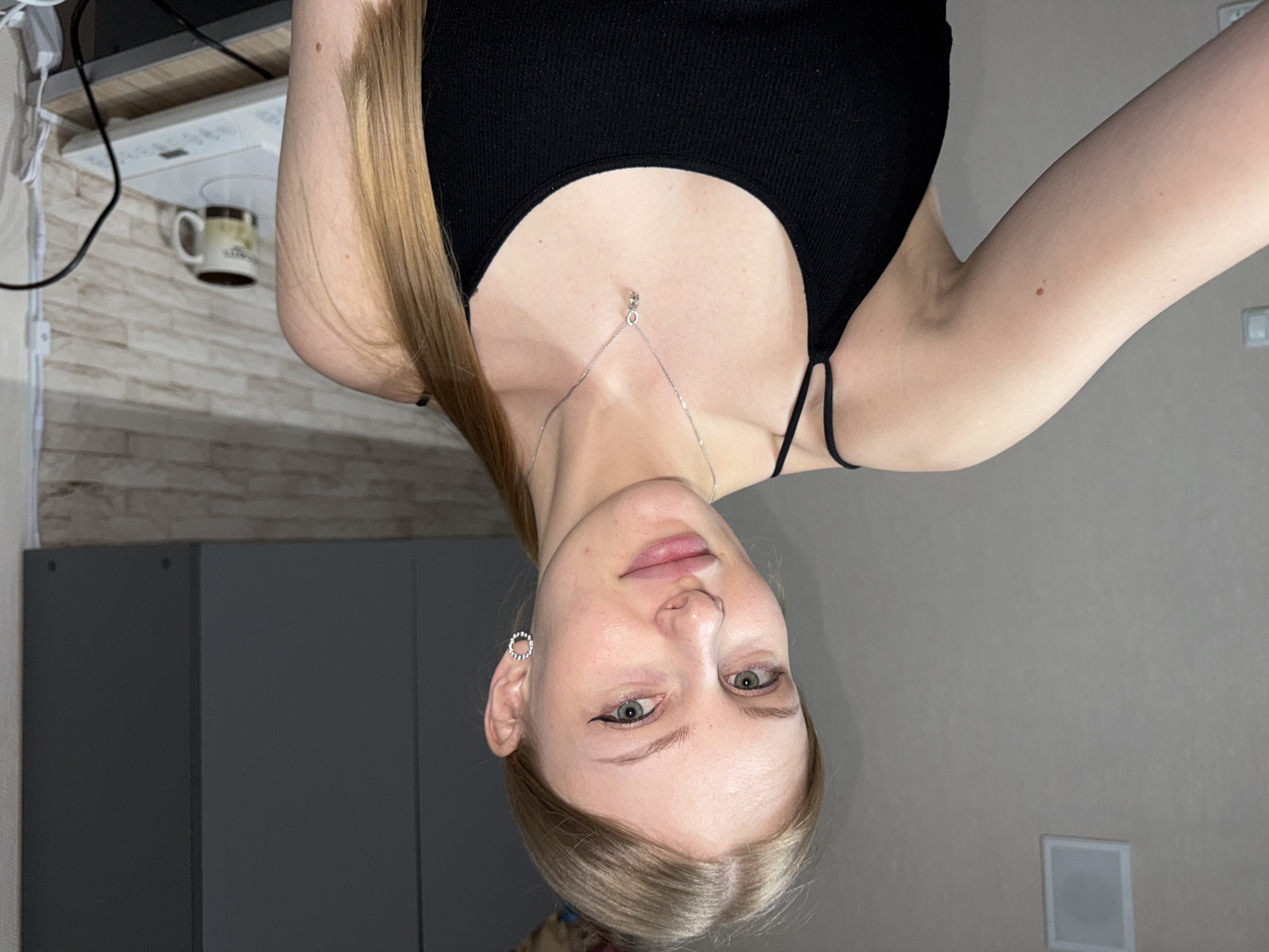 A woman is posing for a selfie. She is wearing a black top and a silver necklace. The photo was taken in an interior setting, most likely a home. A cabinet, a wall, and a mug are visible in the background. It is impossible to confirm the location is in Perm, Russia, without additional information.