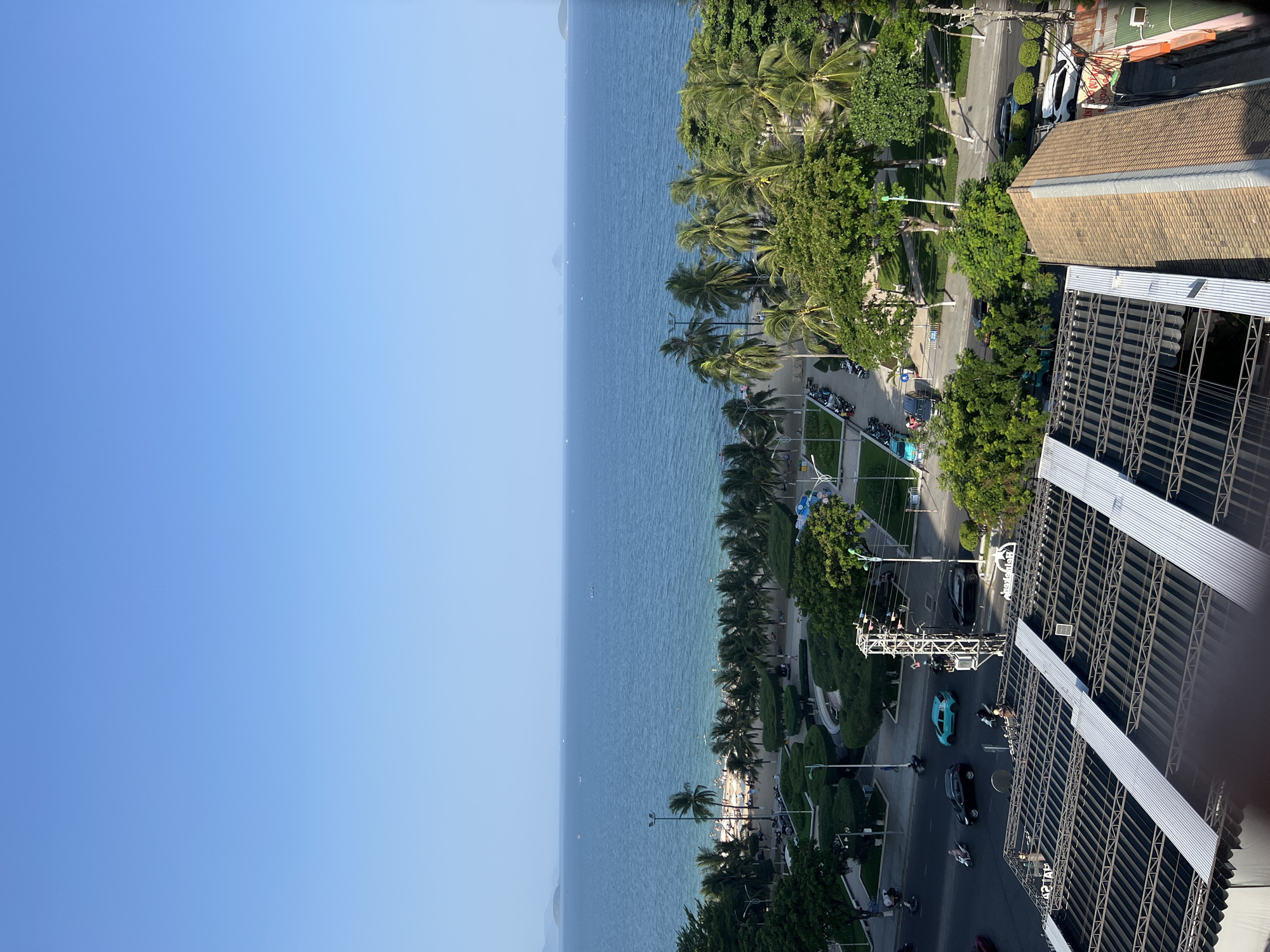 This is an aerial shot of a coastal city, likely Nha Trang, Vietnam, on a clear, sunny day. The foreground features the slanted, corrugated metal roof of a building. Beyond this, a paved road runs parallel to a beach lined with palm trees. The beach is populated with many people sunbathing and swimming in the calm, blue ocean. Several small boats are visible in the distance on the water, and a faint, mountainous landmass can be seen on the horizon. The road is moderately busy with cars and motorcycles. The scene appears tranquil and depicts a typical day at a popular beachside location.