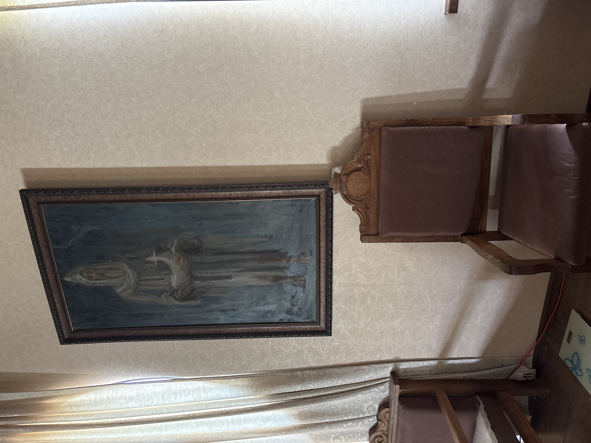 The image depicts an interior room in Gudauta, Abkhazia, featuring a framed painting centered on a light beige wall with a textured, mottled pattern. The painting, presented in a dark, ornate wooden frame, shows a standing male figure with an aged face, long beard, and a hooded or cowled head, wearing a long, light-colored robe. He holds a white bird, resembling a dove or goose, in both hands against a muted blue-grey background, possibly suggesting a forest or desolate setting.
To the left of the painting, light-colored, possibly cream or golden, curtains with a subtle patterned design are drawn back, revealing a window opening from which natural light enters the room. A dark wooden furniture piece, possibly a chair armrest with carved details, is partially visible at the bottom left, adjacent to the curtains.
To the right of the painting and below it, a dark wooden chair with a high back and dark brown, possibly leather, upholstery is positioned, facing slightly towards the left. This chair also features carved wooden elements on its backrest. The floor is composed of dark reddish-brown wooden planks. A red electrical cord runs along the base of the wall near the floor on the left side, and a light-colored mat or paper with blue butterfly outlines is visible on the floor in the lower left quadrant. Shadows are cast on the right wall, indicating light originating from the left.