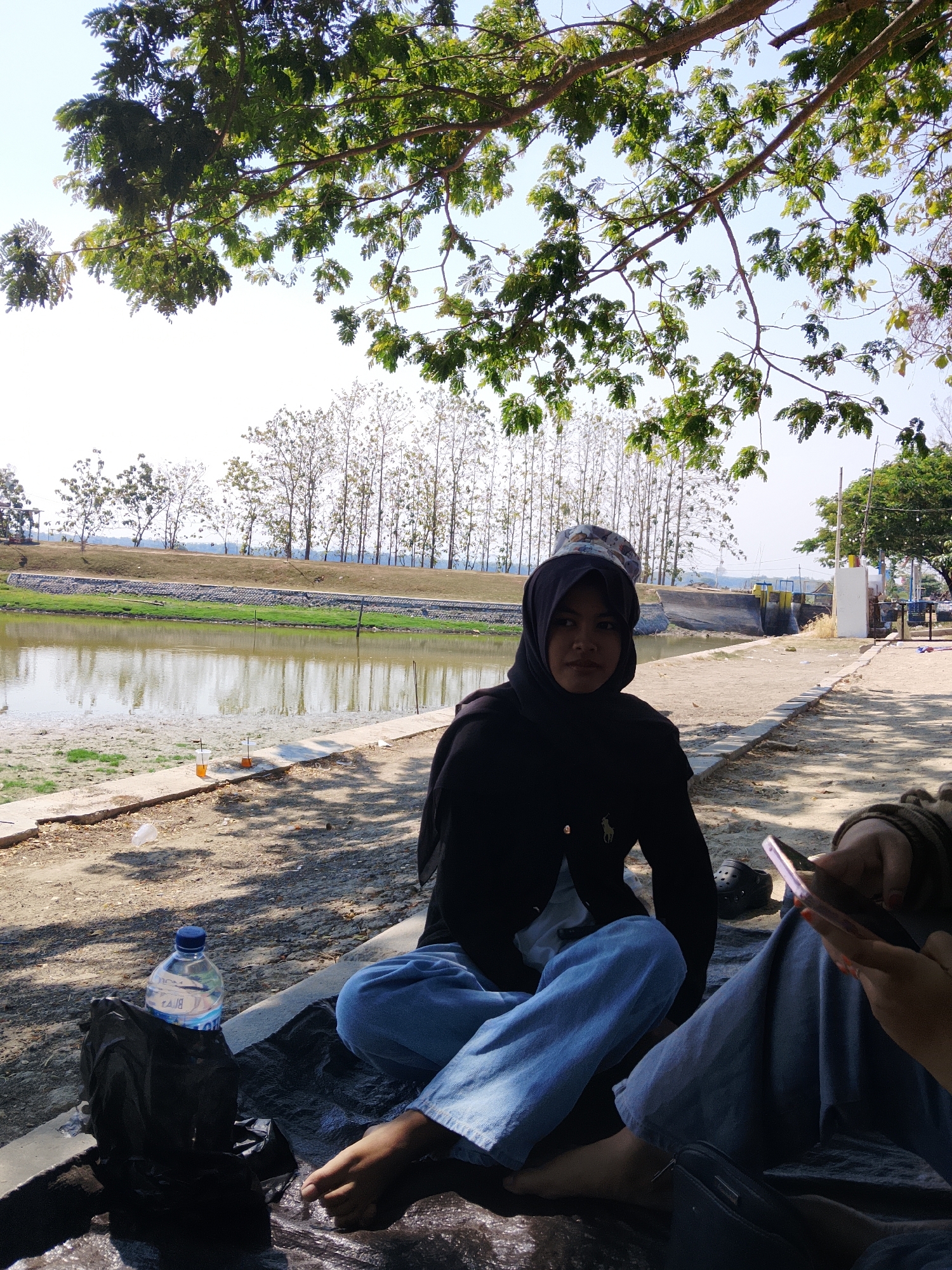 In Ngadrin, Indonesia, on a sunny day, two individuals are seated outdoors by a body of water. One person, wearing a black hijab and a black jacket with light blue jeans, is facing the camera. They have a bucket hat on and their bare feet are visible. Next to them is a black plastic bag and a plastic water bottle. The other individual, to the right, is wearing light blue pants and is holding a smartphone. They are seen from the waist down. The environment features a calm body of water with reflections of trees and the sky. A pathway runs along the edge of the water, with sparse vegetation and a line of trees in the background. The ground is sandy and uneven, with some shadows cast by overhead foliage.