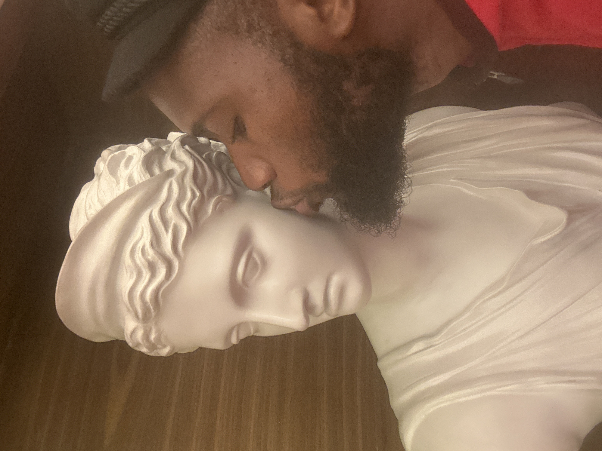 A close-up shot depicts a man with a beard and a black hat leaning towards a white marble bust of a person with Greco-Roman features, appearing to kiss it on the forehead. The scene is indoors, with a dark background suggestive of a room. The image appears rotated slightly. The location in Itirin, Nigeria cannot be confirmed based solely on visual information.