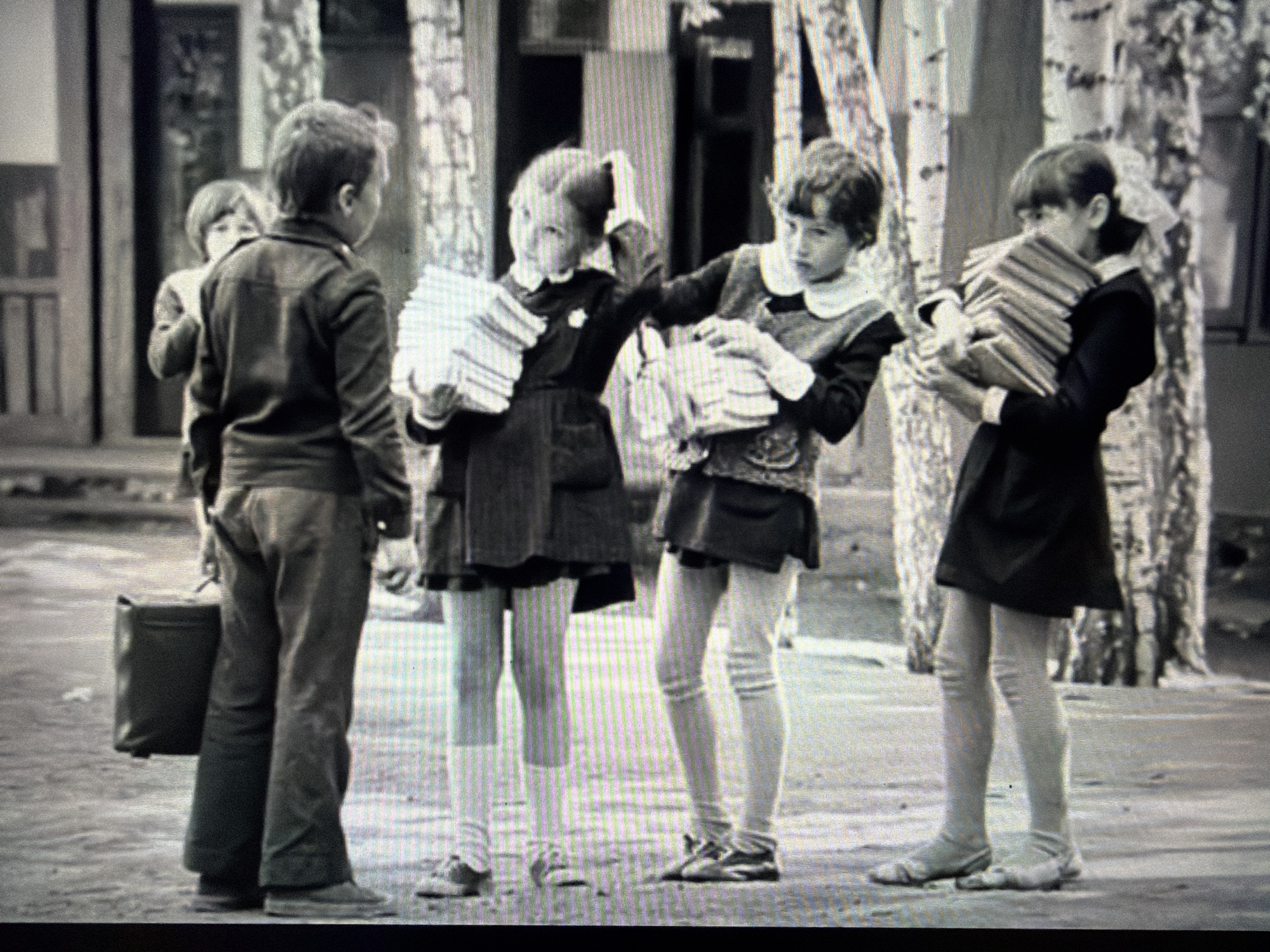 This is a black and white, medium-shot photograph of four children standing in what appears to be an outdoor setting, possibly near a school. The children are dressed in dark clothing, and some are holding stacks of books. The child on the far left is a boy wearing trousers and a jacket, carrying a dark briefcase. To his right is a girl wearing a dark dress and white tights, holding a stack of books. Next to her is another girl in a similar outfit, also holding books. On the far right is a girl carrying a large stack of books. The background shows trees with white bark, possibly birch trees, and a building structure. The lighting suggests it might be daytime. The children's expressions are not clearly discernible due to the resolution and distance. There is no visible text in the image.