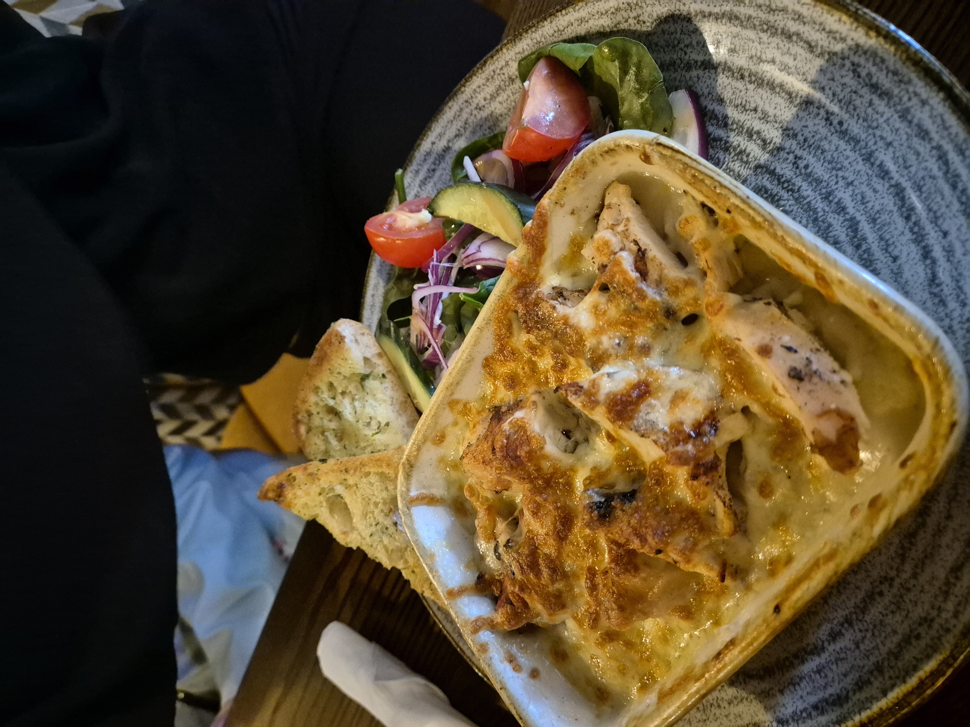 A close-up, high-angle photograph captures a prepared meal served on a speckled grey and brown ceramic plate, likely in a dining setting in Needham Market, United Kingdom. The central focus is a rectangular white baking dish containing a gratin or baked dish, possibly chicken or pasta, with a thick, golden-brown layer of melted, bubbling cheese on top, indicating it was recently baked.
Accompanying the main dish is a fresh side salad, neatly arranged on the plate. It features vibrant green spinach or lettuce leaves, bright red tomato wedges, slices of green cucumber, and delicate slivers of purple red onion. Two pieces of toasted bread, resembling garlic bread, are positioned next to the gratin.
In the upper left background, a person wearing a black top or hoodie is partially visible and out of focus, suggesting they are the diner. Other elements in the blurred background include a patterned fabric, possibly upholstery, and a light blue object. The warm, slightly dim lighting suggests an indoor setting, potentially during the evening. No visible text is present in the image. The scene depicts a casual meal being presented for consumption.