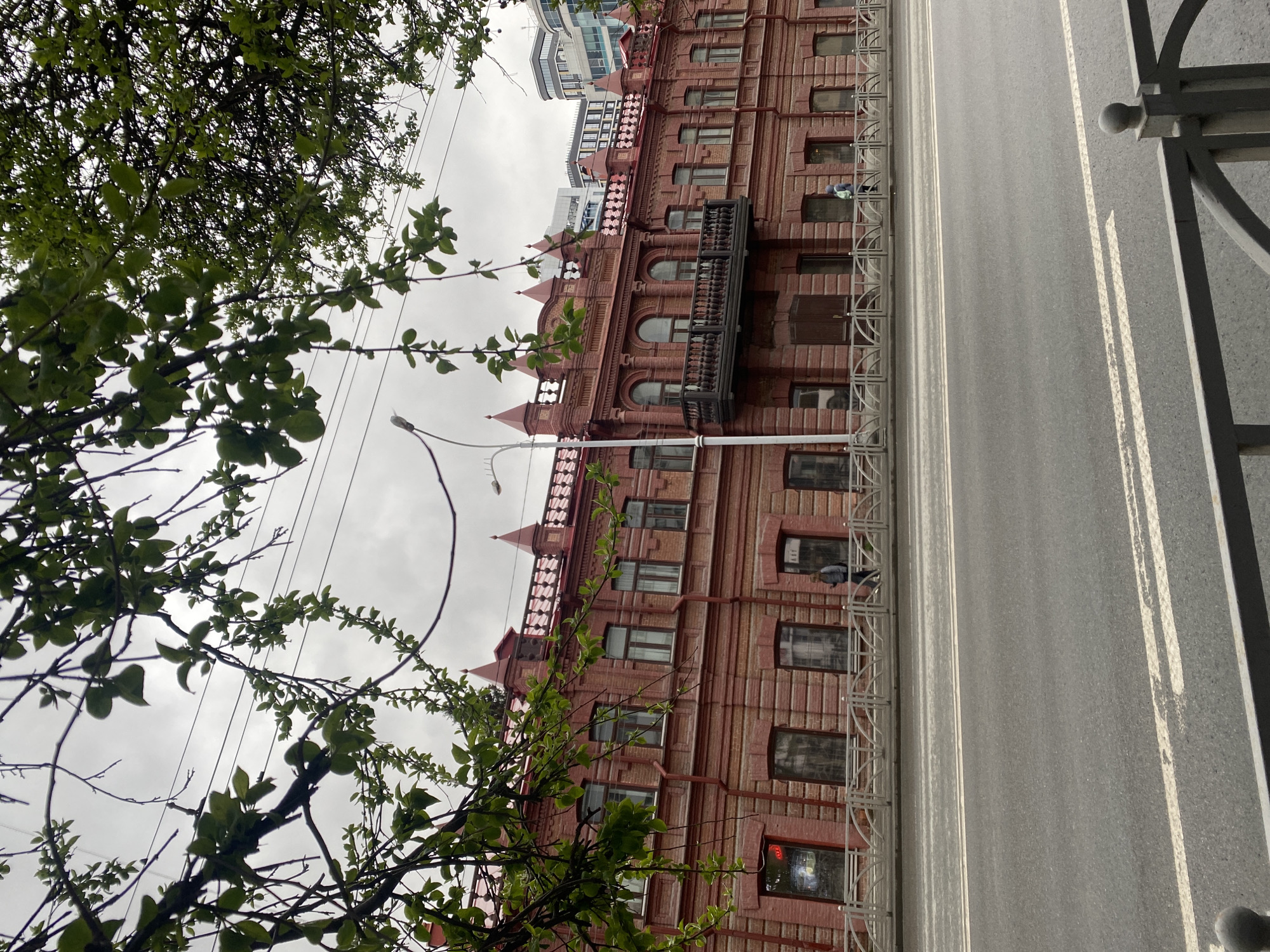 The image displays an urban street scene captured in a vertical orientation, as if rotated 90 degrees clockwise from a standard landscape photograph.
In the foreground, lush green tree branches and foliage extend from the left and top edges, partially framing the scene. Overhead, multiple power lines crisscross against a uniformly overcast, grey sky.
The central subject is a multi-story, reddish-brown brick building situated in the center-left. This structure exhibits classic architectural features, including numerous rectangular and arched windows with dark frames, decorative cornices, and small pointed finials along its roofline. A prominent dark metal balcony with an ornate railing projects from an upper floor. Several air conditioning units are visible beneath some windows. A red glowing sign, with illegible text, is present in a ground-floor window. Behind and above this historic building, modern, taller buildings with lighter facades and reflective windows can be seen. A street light pole is visible near the bottom left of the brick building.
A metal pedestrian fence with a repeating curved design runs along the sidewalk, separating it from the road. On the sidewalk, one person wearing a light-colored top or hoodie stands facing away from the camera, near the fence line. Another individual is faintly discernible within an upper-story window of the brick building.
The paved road surface occupies the center-right of the image, running vertically. It features distinct white double solid and single dashed lane markings. On the far right edge, a section of a decorative metal fence with spherical adornments borders the road.
The scene depicts an ordinary urban moment on what appears to be a cloudy day, given the diffused lighting and absence of strong shadows. The fresh green leaves suggest spring or early summer. The blend of older brick architecture with newer buildings in the background indicates an evolving city landscape. No specific event or strong emotions are conveyed, portraying a calm, everyday street view.