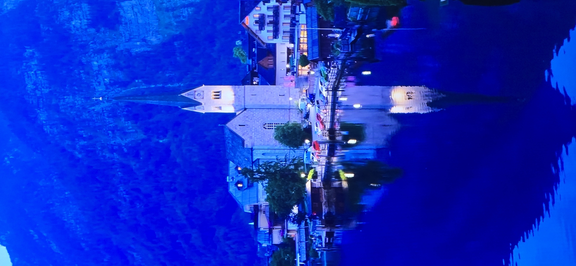 This is a photograph of a serene waterfront scene at dusk or twilight, with the entire image appearing rotated 90 degrees counter-clockwise. The dominant color is a deep, rich blue, indicative of the evening sky and the dark, reflective water.
The scene features a charming village nestled along the water's edge. Prominently in the foreground, a substantial stone building, possibly a church, with a steepled roof and arched windows, stands out. Adjacent to it, other buildings with illuminated windows suggest a lively town center. These structures are reflected in the calm water, creating a mirror image that blurs the lines between reality and reflection. A narrow pier or walkway extends into the water, lined with lights and several small boats or kayaks tied up. Trees with dark foliage add a touch of nature to the urban setting.
The time of day is clearly evening, with artificial lights beginning to illuminate the buildings and the waterfront, contrasting with the fading natural light. The weather appears calm, with the water being still enough to produce clear reflections. The location is suggested to be Drummondville, Canada, though specific landmarks that confirm this are not immediately apparent. The overall mood is peaceful and tranquil, evoking a sense of quiet beauty. No visible text is present in the image.
