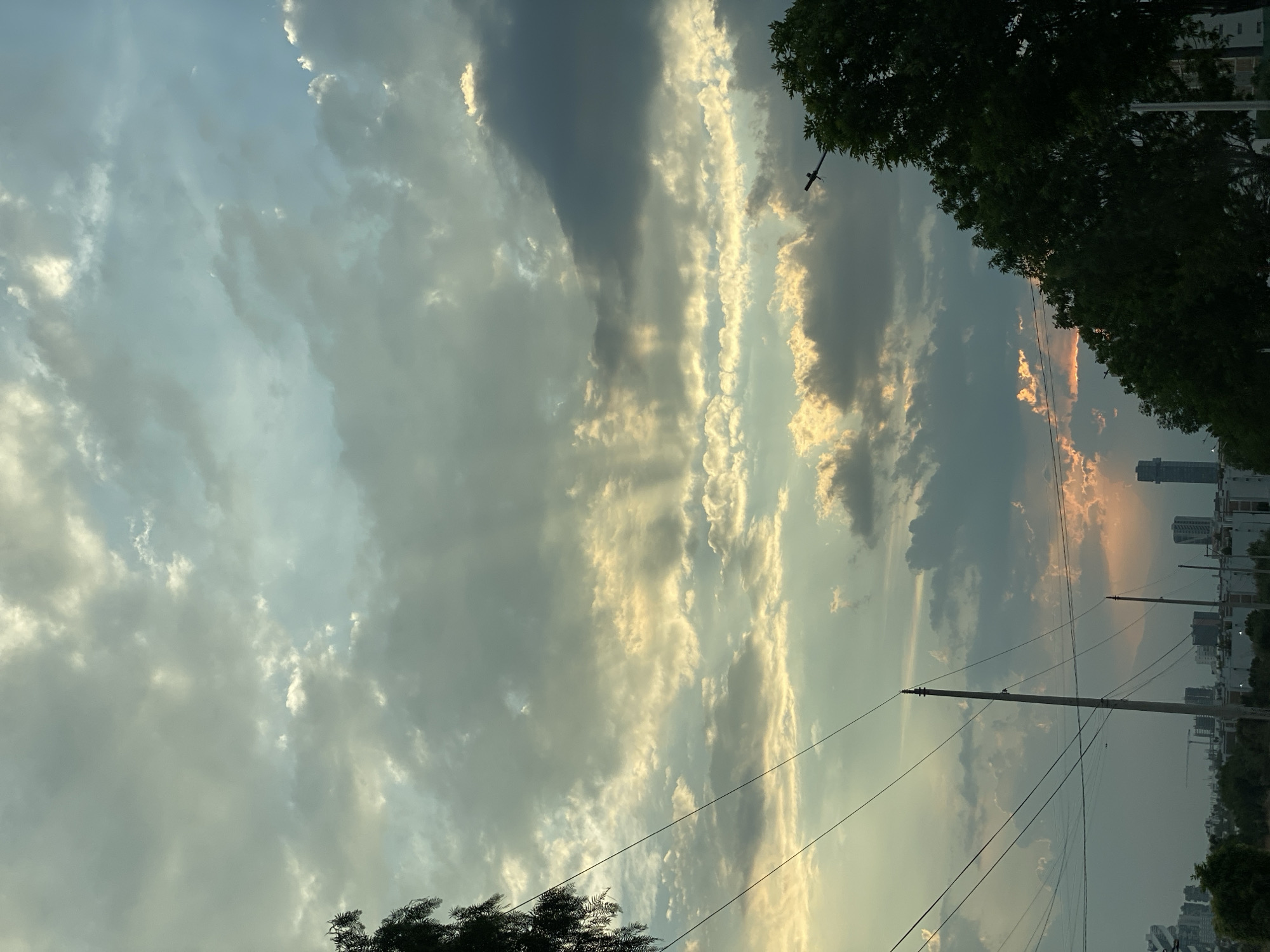 The image depicts a partly cloudy sky above a city. The clouds vary in color from dark gray to bright white, with some golden tones suggesting the presence of sunlight. In the lower right corner, there are buildings and trees, indicative of an urban environment. Utility poles and power lines are visible, cutting across the sky. The location appears to be within a city, but the specific city is not identifiable from the image.