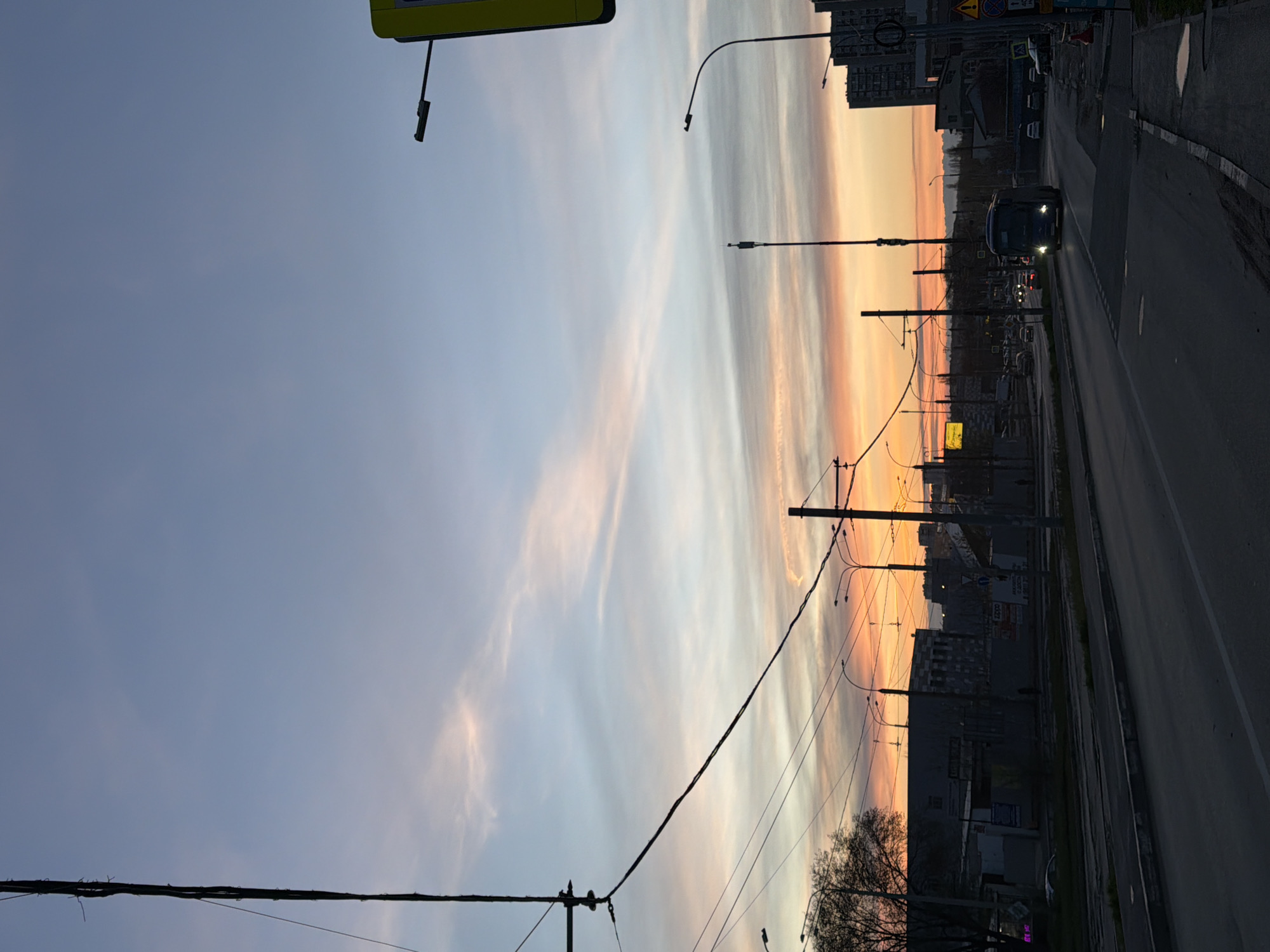 The image depicts a street scene in Rostov-on-Don, Russia at sunset. The sky is a blend of blue and orange, reflecting the fading sunlight. A road runs along the right side of the frame, and a vehicle with headlights on is visible on the road. Buildings and utility poles are silhouetted against the colorful sky. Wires stretch across the sky, connecting the poles.