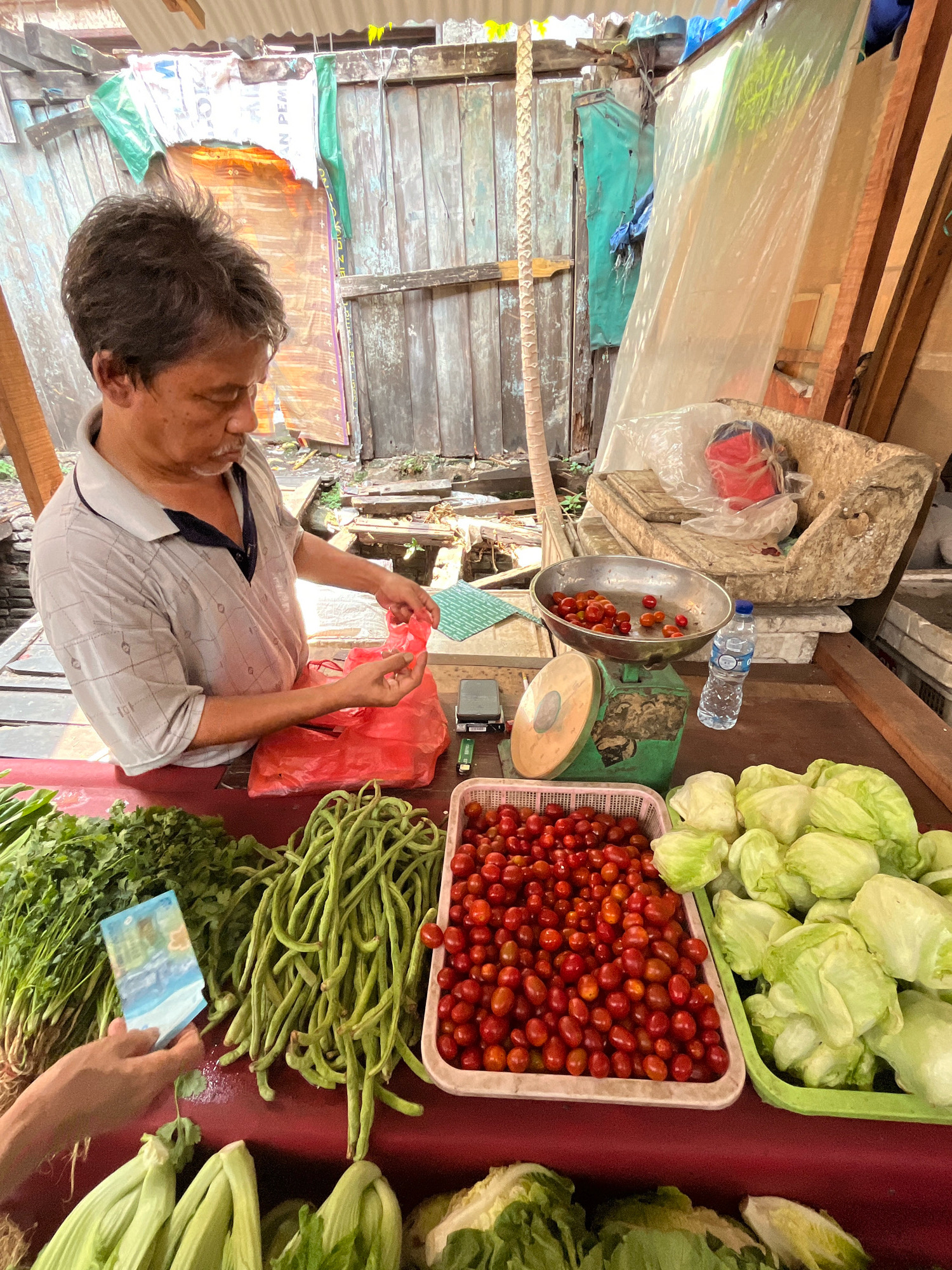 A person is standing behind a market stall in an outdoor setting, preparing to bag vegetables. The stall is laden with fresh produce including bunches of green onions, long beans, cherry tomatoes in a basket, and heads of lettuce. In the background, there are wooden structures, tarps, and what appears to be old scaffolding. A mechanical scale with a bowl of cherry tomatoes on it is visible on the counter. A person's hand is holding a banknote, possibly for a purchase. The environment suggests a traditional market.