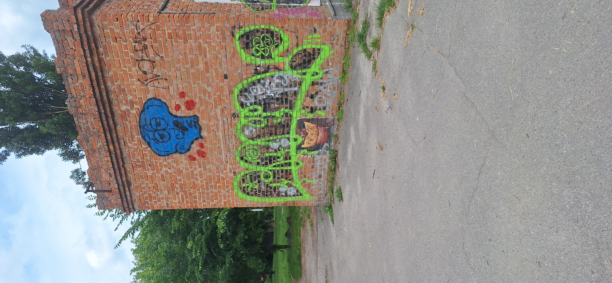 A brick building stands to the right, its upper edge adorned with decorative brickwork and a metal fixture. The building's surface is covered in graffiti, with a large blue cartoon character on the upper portion and bright green lettering across the lower half. A small, yellow, cheese-like object with a cartoon face is depicted below the green lettering. To the left of the building, lush green trees and grass are visible, suggesting an outdoor, possibly urban or park-like setting. The ground in front of the building is asphalt. This image was taken in Bila Tserkva, Ukraine.