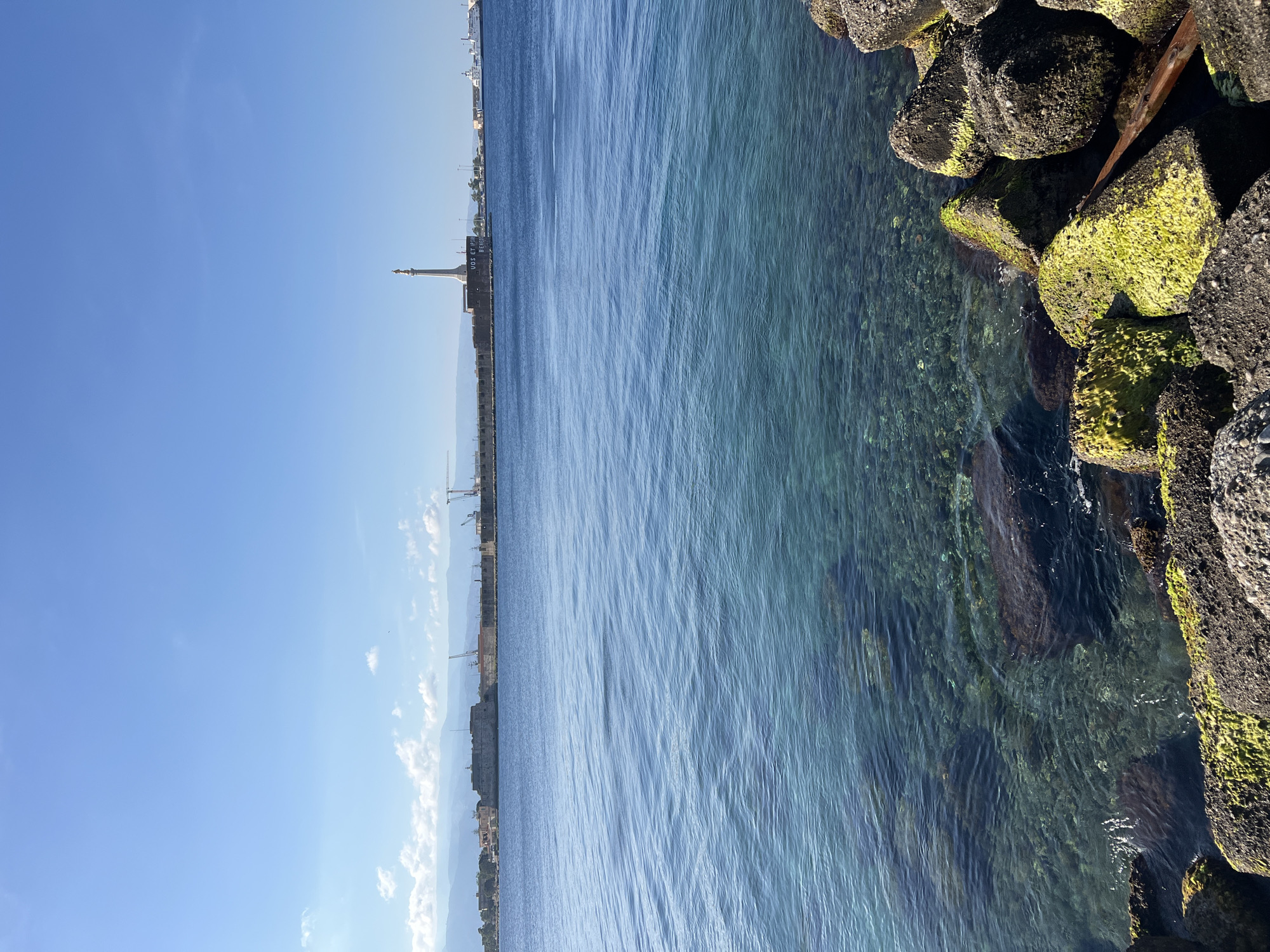 The image depicts a sunlit coastal panorama in Messina, Italy, overlooking the city and its harbor. In the immediate foreground, dark grey, angular concrete breakwater rocks, extensively covered in vibrant green moss and algae, line the bottom right edge. A piece of dark wooden debris is lodged among these rocks. The water in the mid-ground is remarkably clear, displaying a gradient of deep blue to turquoise, allowing the rocky seabed and submerged features to be distinctly visible beneath the surface. Gentle ripples texture the water.
Across the water, the distant coastline reveals the city of Messina. A prominent white column, the Madonna della Lettera monument, stands tall on a fortified base at the harbor entrance. A partial inscription, "VOS ET IPSAM BENEDICIMUS," is visible on its pedestal. To the left of the monument, a large, dark, fortified structure or wall forms part of the port defenses. Further along the distant shore, industrial port infrastructure is present, including multiple yellow construction cranes, various buildings, and a large white vessel, possibly a ferry, docked at a terminal. In the far background, a range of hazy, undulating mountains and hills extends across the horizon. The sky is a clear, bright blue with a few scattered white cumulus clouds. No individuals are discernible in the scene.