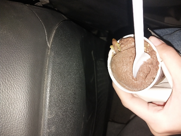 A hand holds a small white cup of chocolate ice cream with bits of nuts. A white plastic spoon is plunged into the ice cream. The background features a dark grey leather or vinyl car seat. The ice cream appears to be sitting in a car, possibly being enjoyed during a ride.