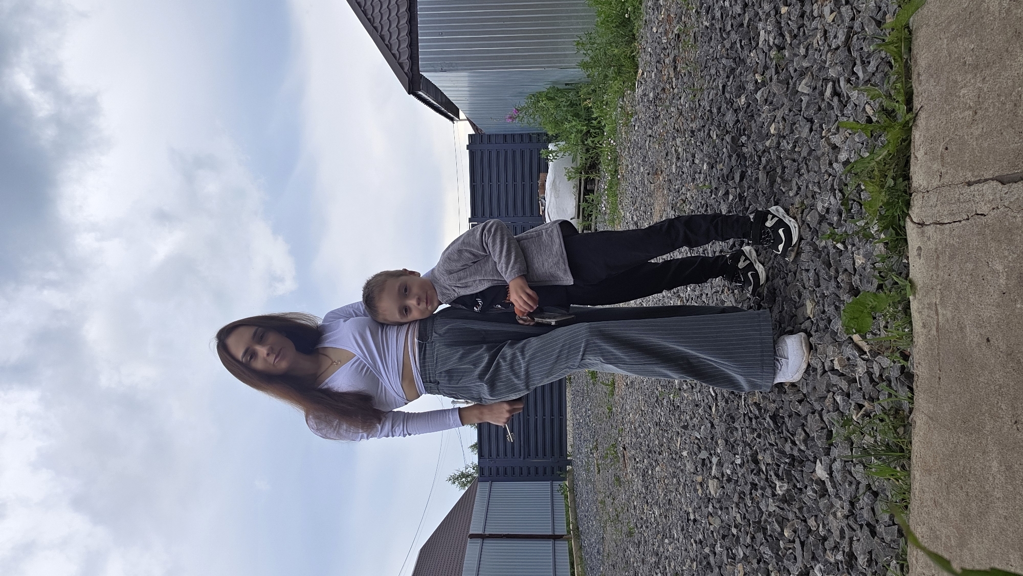 A young woman and a young boy stand close together outdoors in a yard. The woman is taller and stands on the left, wearing a white crop top and grey pinstriped wide-leg pants. She has long brown hair and is looking towards the camera. The boy, on the right, is wearing a grey zip-up jacket over a dark t-shirt and black pants. He has short brown hair and is also looking at the camera, with his left arm around the woman's waist. They are standing on a gravel ground. Behind them is a dark fence and a corrugated metal building. The sky above is cloudy and grey.