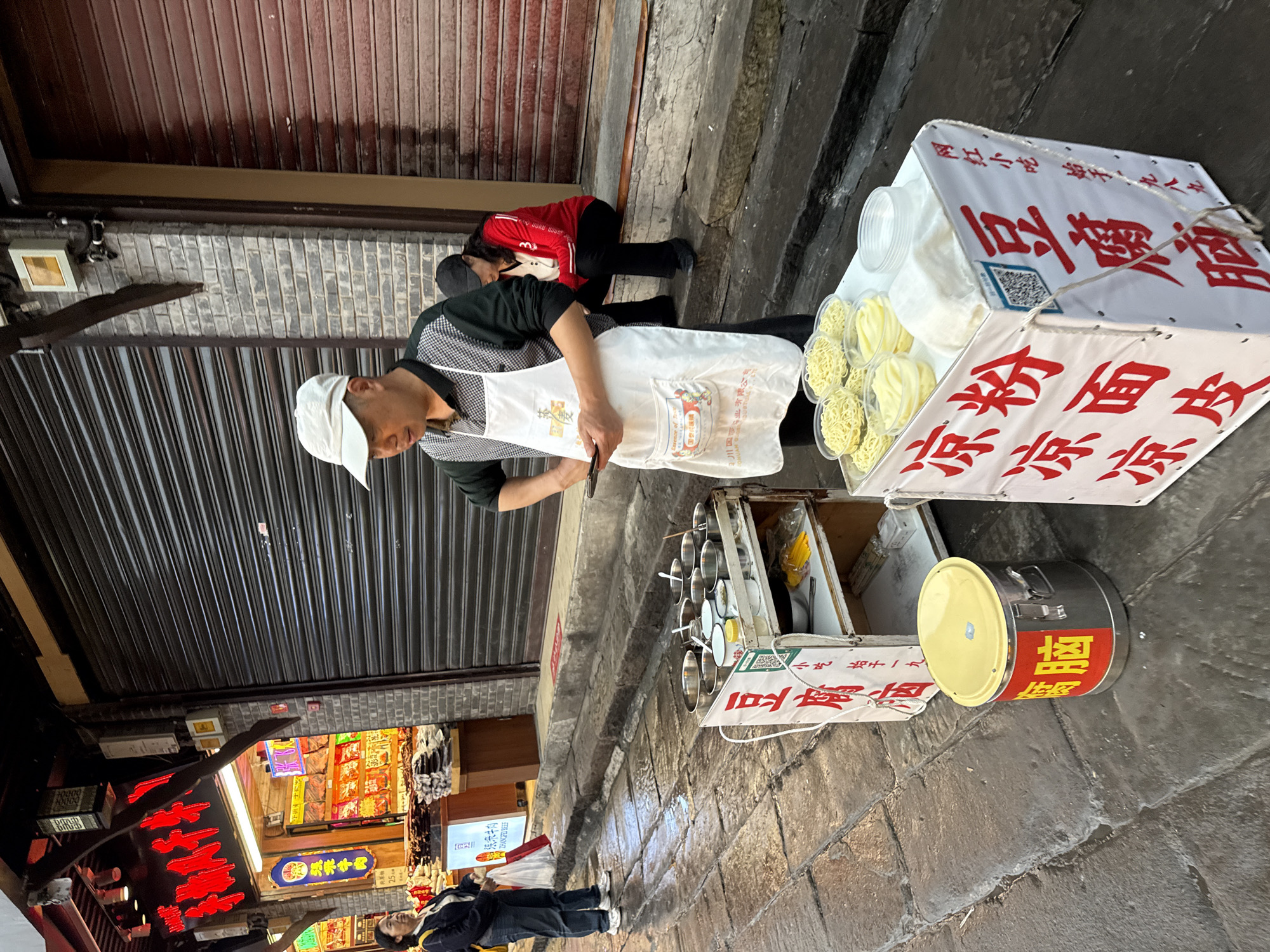 A street vendor in Shapingba District, China, wearing a white apron and a white baseball cap, stands behind a food cart on a paved street. The cart displays containers of noodles and a container labeled "豆腐脑" (Tofu Pudding) in Chinese. A woman in red bends down next to the cart. In the background, a store with illuminated signs is visible. Other pedestrians walk past the vendor and shop.