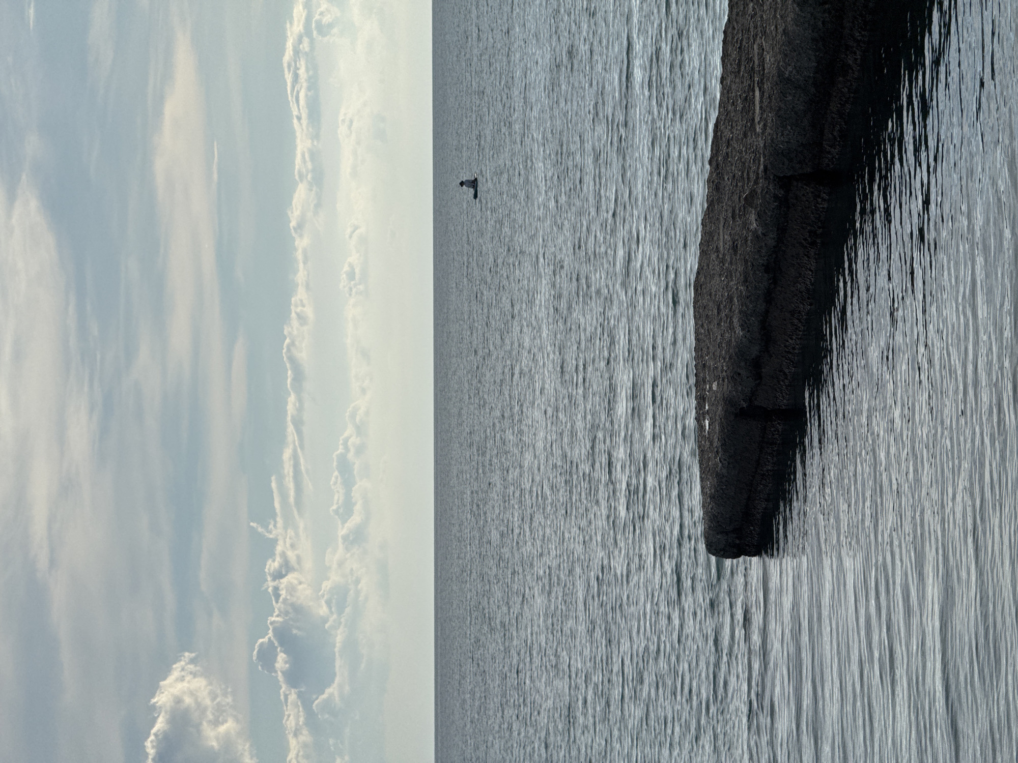 This image depicts a calm body of water, likely the Black Sea, with a wide expanse of water stretching to the horizon. In the foreground, a dark, rectangular concrete structure juts out into the water, partially submerged. The surface of the water is rippled, suggesting a gentle movement. In the distance, a single person is visible on a paddleboard, silhouetted against the horizon. The sky above is partly cloudy, with soft, diffused light creating a serene atmosphere. The scene evokes a sense of tranquility and solitude. The location is not specified in the image, but given the context of Sochi, Russia, this could be a typical coastal view from its shoreline.