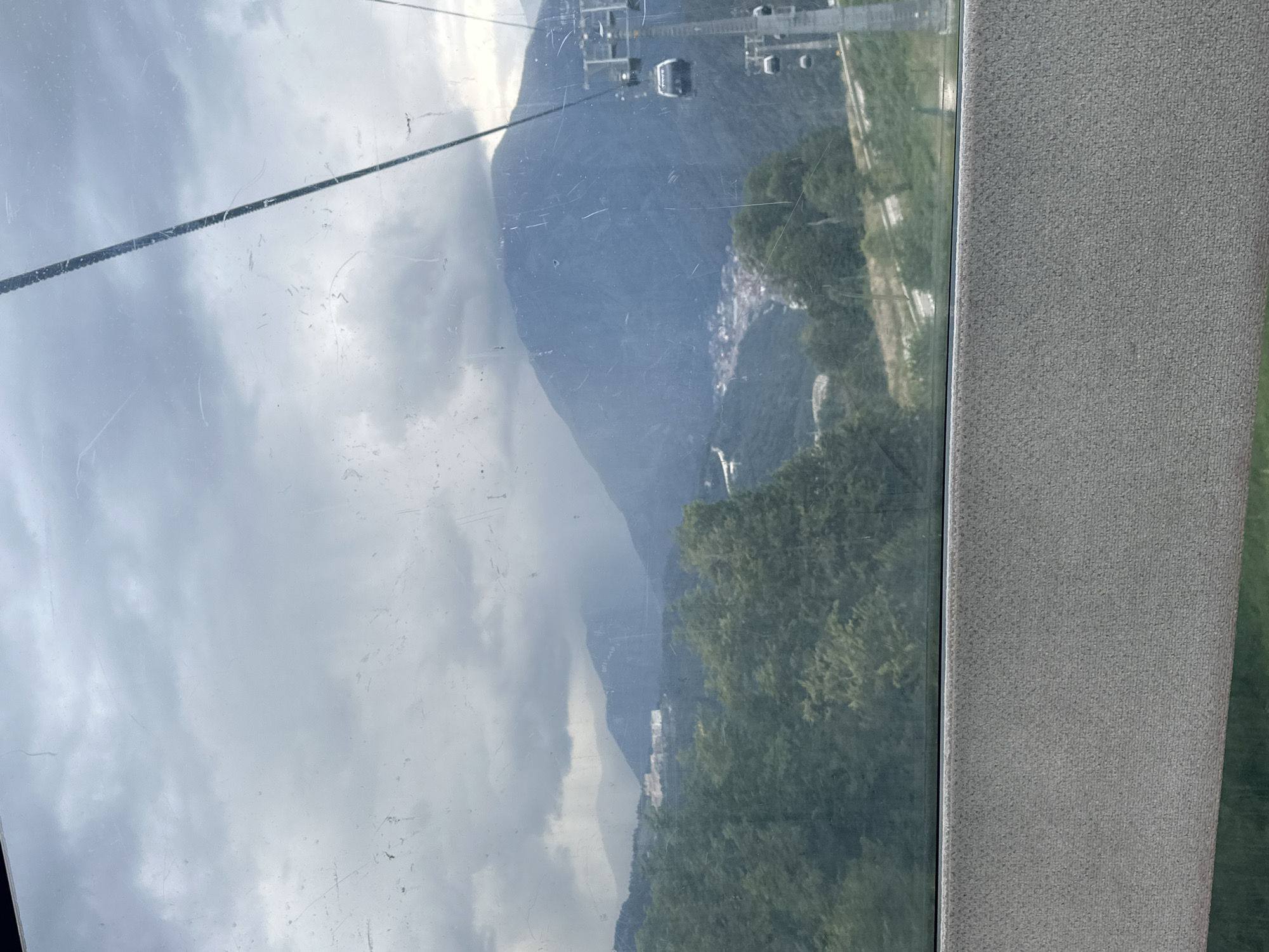 This image depicts a view from a cable car in Estosadok, Russia. The cable car itself is partially visible, with one car and a support tower on the right side of the frame. The background features a lush, green mountain landscape under a cloudy sky. In the mid-ground, a cluster of buildings, likely a village or resort, is nestled on a mountainside. The window of the cable car is streaked and scratched, obscuring some of the finer details of the scene. The foreground shows the textured interior of the cable car, with a grey, carpeted surface. The overall atmosphere is one of natural beauty and a sense of travel.