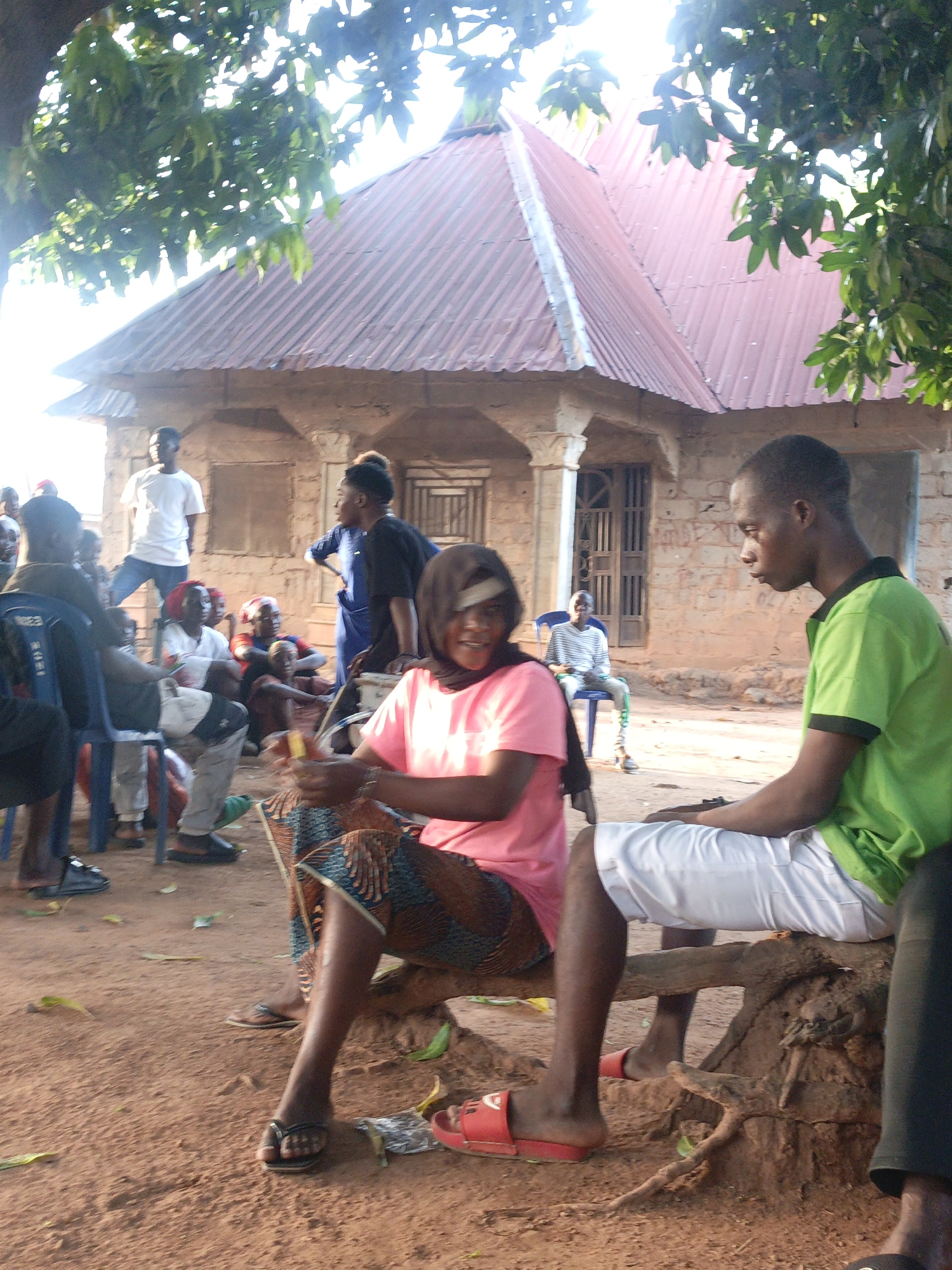 The image depicts an outdoor gathering in Akwanga, Nigeria. A group of people is assembled in front of a single-story building with a red corrugated iron roof. A woman in a pink shirt and patterned skirt sits on a wooden structure, possibly crafting or repairing something with her hands. A man wearing a green shirt and white shorts is seated next to her. Other individuals are seated or standing around them, some on chairs. The ground is reddish-brown dirt, and there are trees providing some shade.