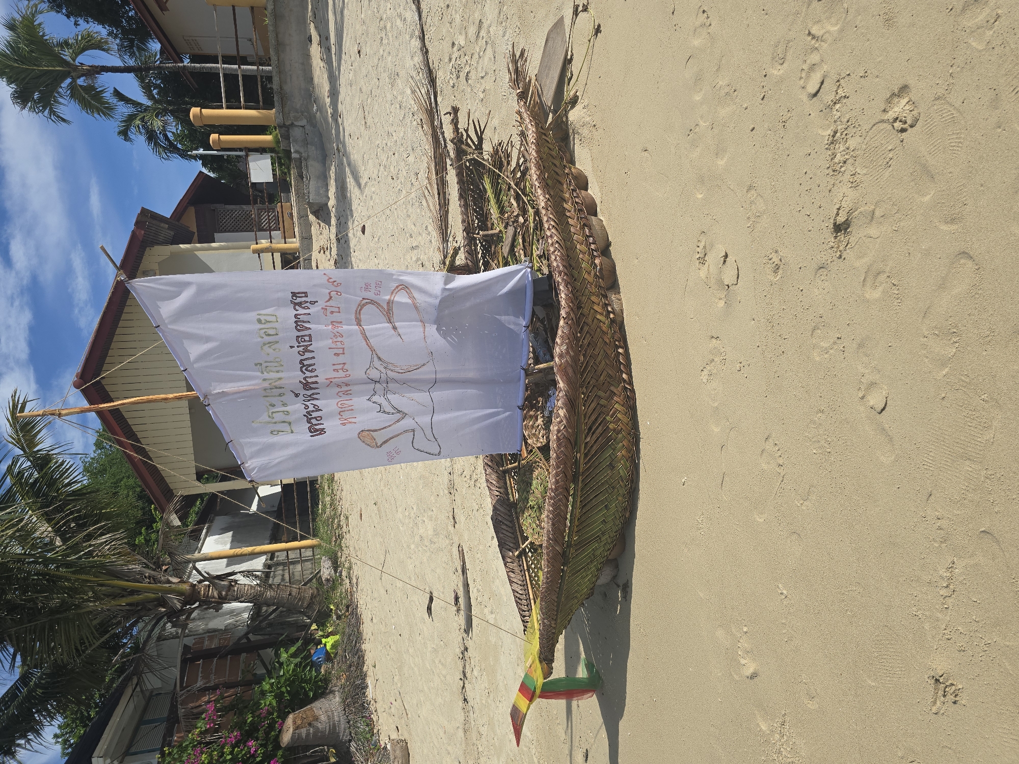 A wide shot shows a handcrafted boat on a sandy beach in Baan Lamai, Thailand. The boat is made of woven palm fronds and appears to be supported by coconut shells. A white sail, attached to a wooden mast, is erected on the boat. The sail has text and a drawing of a person with a heart. Palm trees and buildings line the background, with a clear blue sky overhead. The sand is marked with footprints.